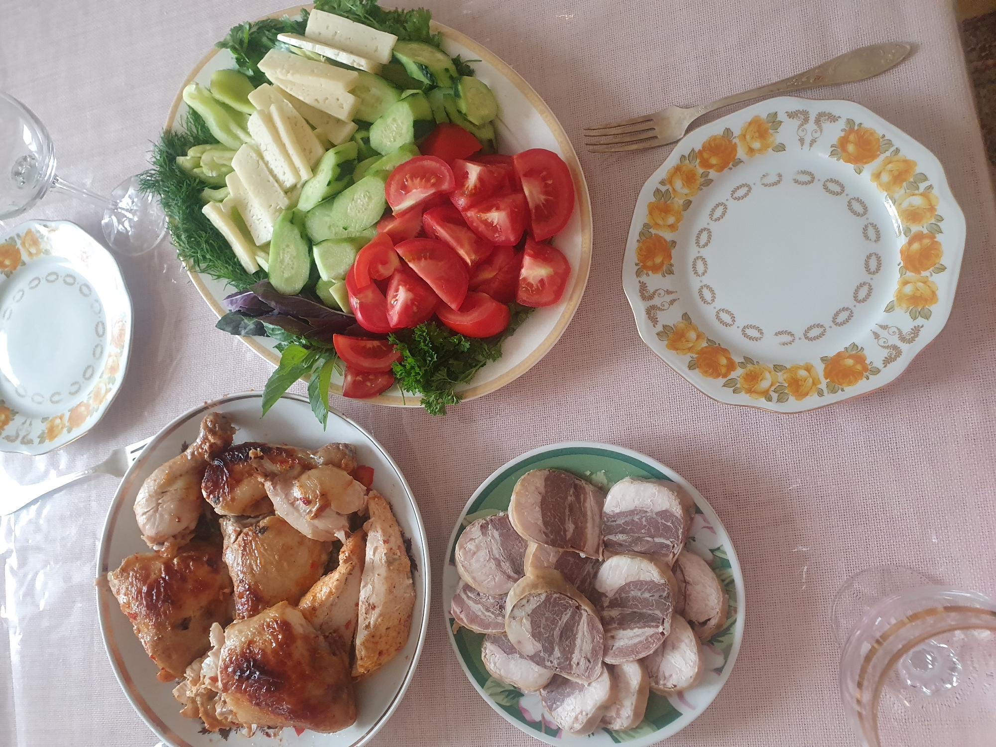 A top-down shot shows a table set for a meal in Almaty, Kazakhstan. The table is covered with a pink, textured tablecloth. Several plates of food are arranged on the table, suggesting a meal is about to be served or has just been consumed.
In the upper left quadrant of the image, a plate of sliced cheese, cucumbers, tomatoes, and herbs is visible. To its right, a decorative plate with a floral pattern sits next to a fork.
Below these, on the left, a plate holds several pieces of roasted chicken, including drumsticks and other parts. To the right of the chicken, a plate displays an assortment of sliced meat rolls, with visible layers of fat and lean meat.
A glass with a golden rim is partially visible in the lower right corner. The overall scene conveys a home-style meal setting, likely a casual gathering or family meal. The lighting suggests daytime. There is no visible text in the image.