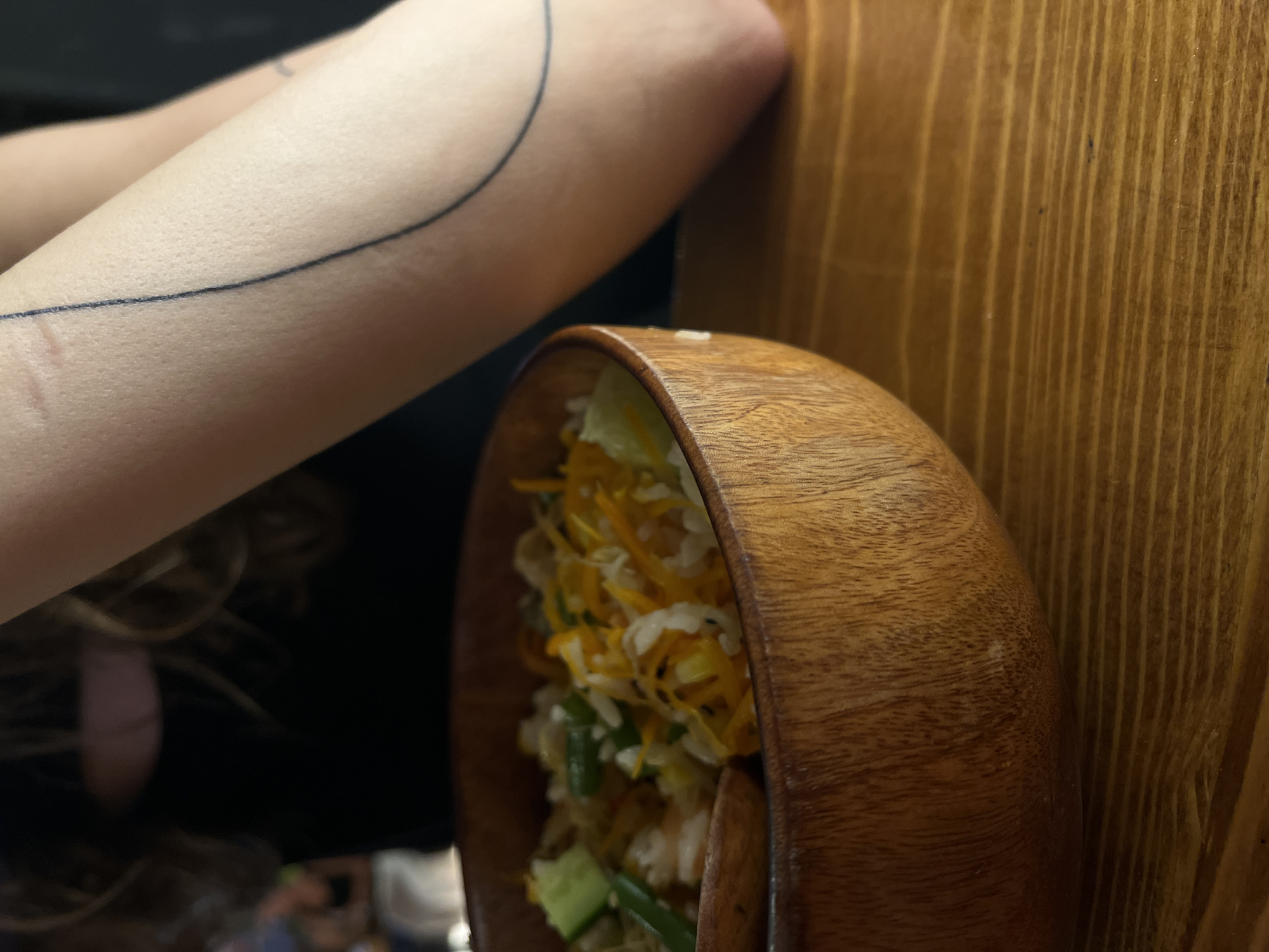 A close-up, slightly angled, dimly lit image shows a wooden bowl containing a salad. The salad appears to contain shredded cabbage, carrots, green beans, and cucumbers. The bowl sits against a wooden wall with vertical grain. To the left, an arm with a black line tattoo is visible. The photo was likely taken indoors, with the subject possibly seated at a table. The city of Đà Nẵng, Vietnam, and the exact location within the city are not explicitly evident from the image.