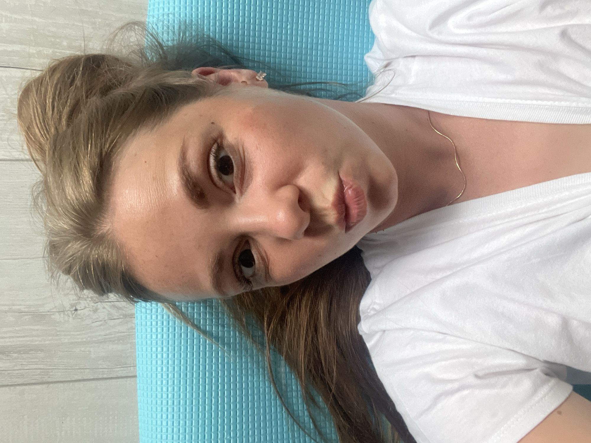 The media file shows a close-up, vertically oriented selfie of a young woman lying down on a textured blue mat. She is wearing a white t-shirt and a delicate gold necklace. Her hair is pulled up loosely, with some strands falling around her face. Her facial expression is relaxed and somewhat playful, with her lips slightly pursed and her gaze directed towards the camera. A small, sparkling earring is visible on her left ear. The background consists of a light-colored, horizontally paneled surface, resembling a wooden wall. The lighting suggests it is daytime. The overall mood is casual and intimate. No visible text is present in the image. The provided location context is Chelyabinsk, Russia.