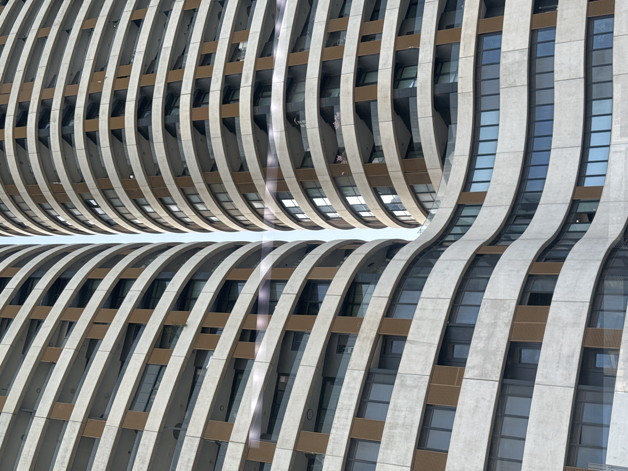 The image shows a close-up of a modern architectural structure, specifically a section of a building in Tel-Aviv, Israel, known as the "David Promenade Residences". The building's facade features a series of vertical, curved concrete ribs that run along its height, creating a distinctive wave-like pattern. These ribs are spaced apart, revealing the building's windows and balconies. The windows are rectangular and span multiple floors. Brown decorative elements are visible between the concrete ribs, adding a horizontal layer to the design. The overall appearance is abstract and emphasizes the building's unique architectural design. The environment appears to be daylight, with even lighting across the facade.