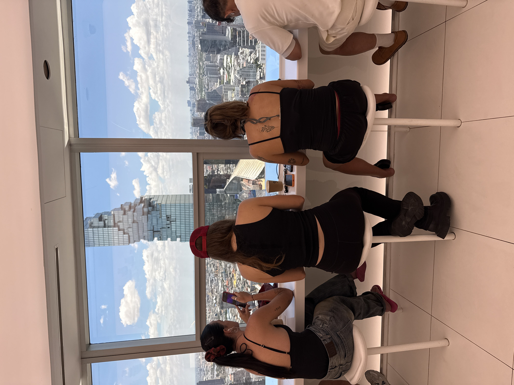 The image shows four people sitting at a white counter looking out a large window. The view outside is of Bangkok, Thailand, with a distinct building known as the MahaNakhon skyscraper visible. The people are seated on white stools at a long counter. One person is wearing a white shirt and brown shoes, while the other three are wearing black tank tops. One of them is also wearing a red cap and another person is holding a cell phone. The setting seems to be an observation deck or a high-rise bar/lounge.