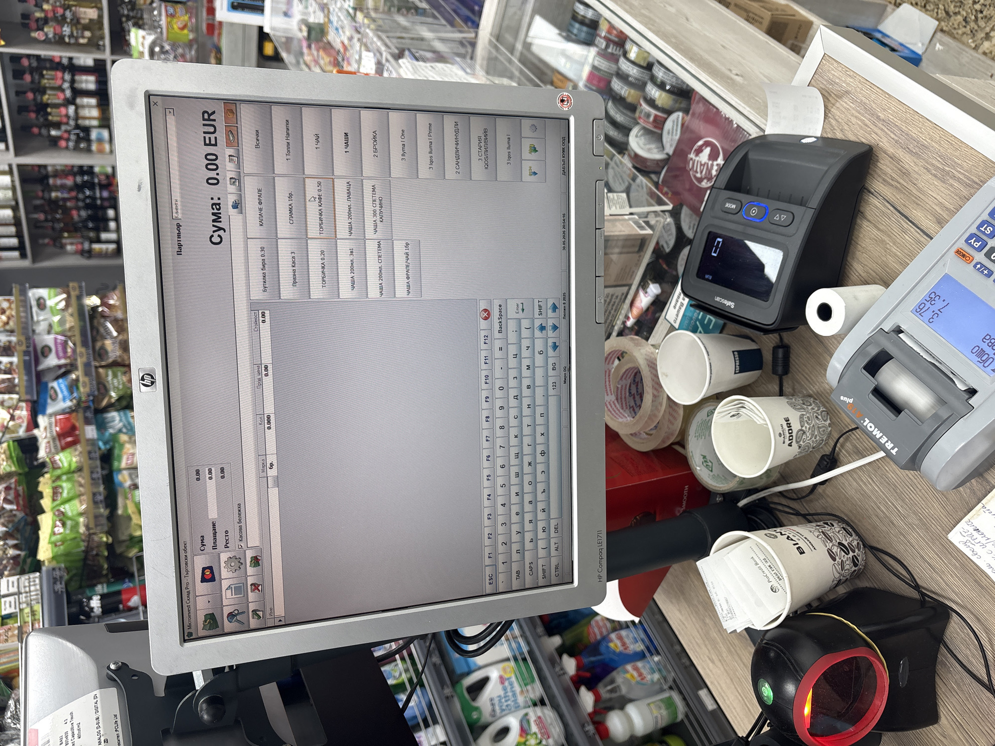 The image shows a cashier's station in a store in Sofia, Bulgaria.
On the counter is a point-of-sale (POS) system with an HP Compaq LE1711 monitor showing the "Microinvest Склад Pro - Търговски обект" software interface with a sales transaction of "0.00 EUR". The interface lists item options in Bulgarian, including "Бутилка бира" (beer bottle), "КАПАЧЕ ФРАПΕ" (frappe lid), "Топли Напитки" (hot drinks), and others.
Next to the monitor are several devices: a Safescan money counter, a Tremol Plus cash register, and a barcode scanner. There are also used paper cups on the counter.
In the background, shelves are visible with various goods, including liquor, snacks, and cleaning supplies.