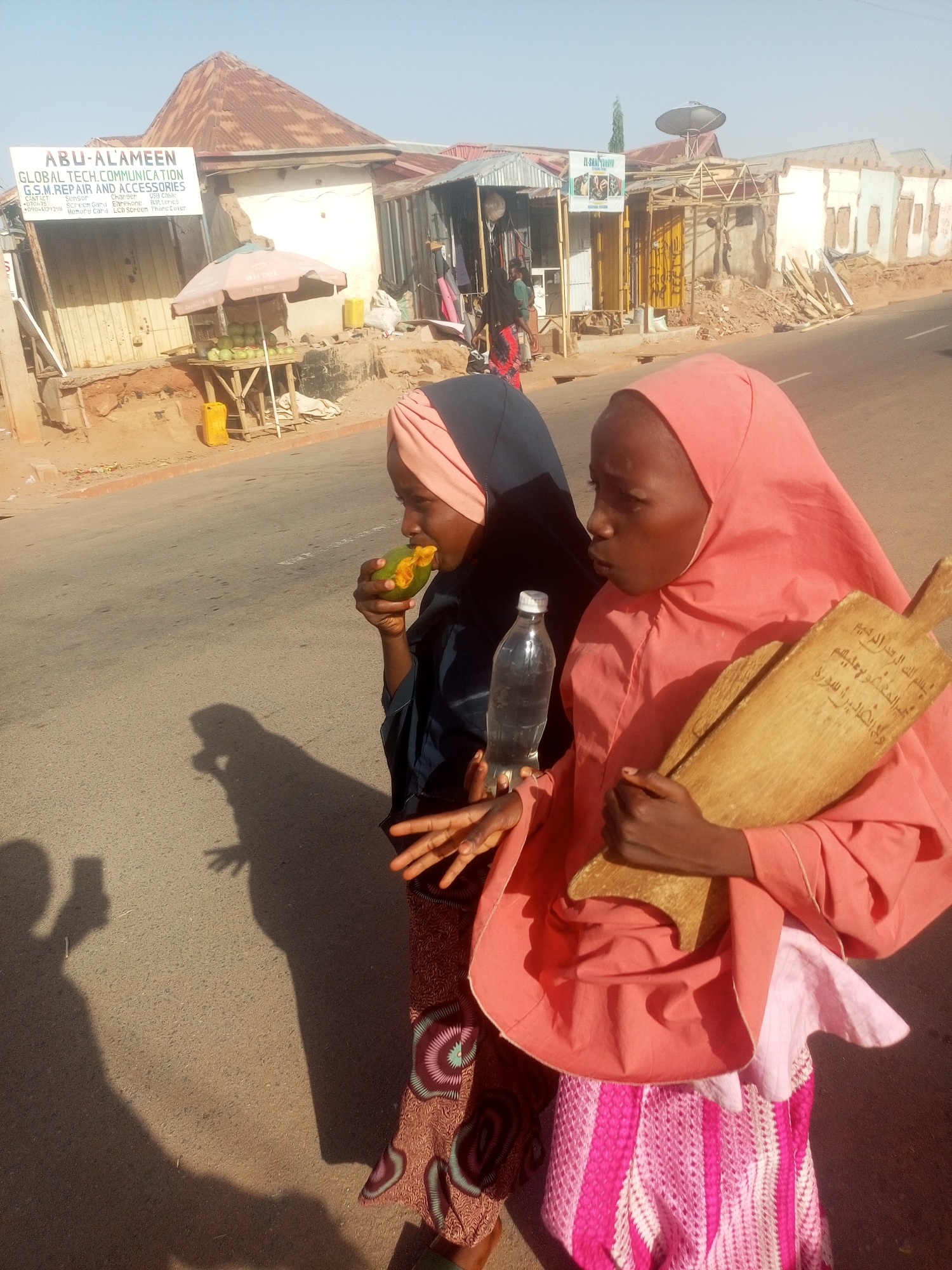 Two young women walk on a paved street in Gombe, Nigeria. The woman on the left wears a black hijab and a patterned skirt and is eating a piece of fruit. The woman on the right wears a pink hijab and skirt and carries a wooden board with Arabic writing. She also holds a clear plastic water bottle. The shadow of the photographer holding a mobile phone to take the picture is visible on the street. Behind the women are businesses with signs in English and Arabic. One sign advertises "ABU-ALAMEEN GLOBAL TECH COMMUNICATION GSM REPAIR AND ACCESSORIES." Another business appears to sell fruit from under a pink umbrella. The street is lined with small shops and buildings. A satellite dish is visible on a nearby building.
