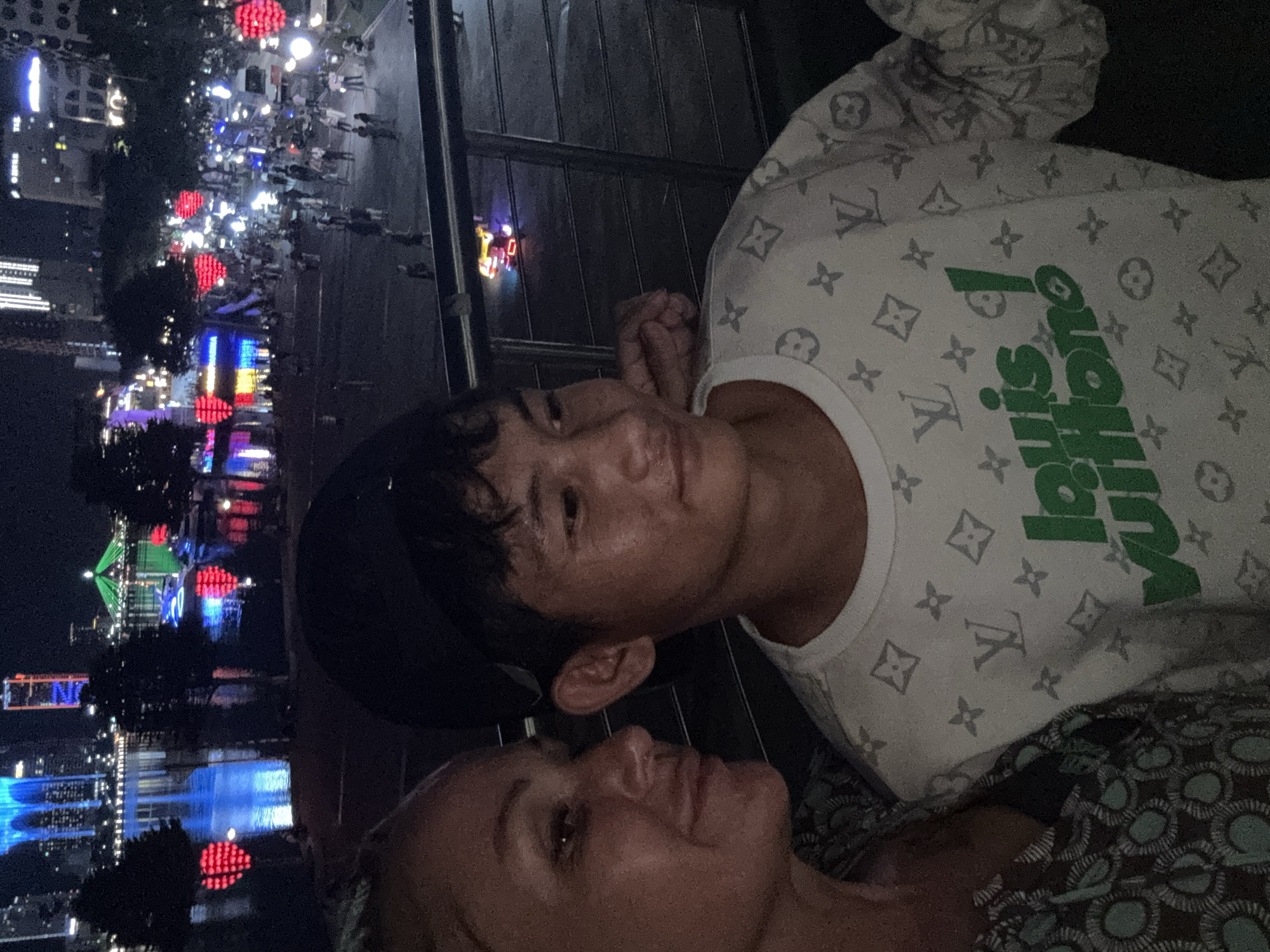 Two people are positioned in the foreground, with one wearing a Louis Vuitton-patterned shirt. The background features a brightly lit urban landscape at night. A body of water reflects the lights, and various structures, including buildings and illuminated objects, are visible. The location appears to be in the city of Đà Nẵng, Vietnam. Crowds of people are also present in the background. The individuals in the foreground appear to be interacting, though the exact nature of their interaction is unclear.