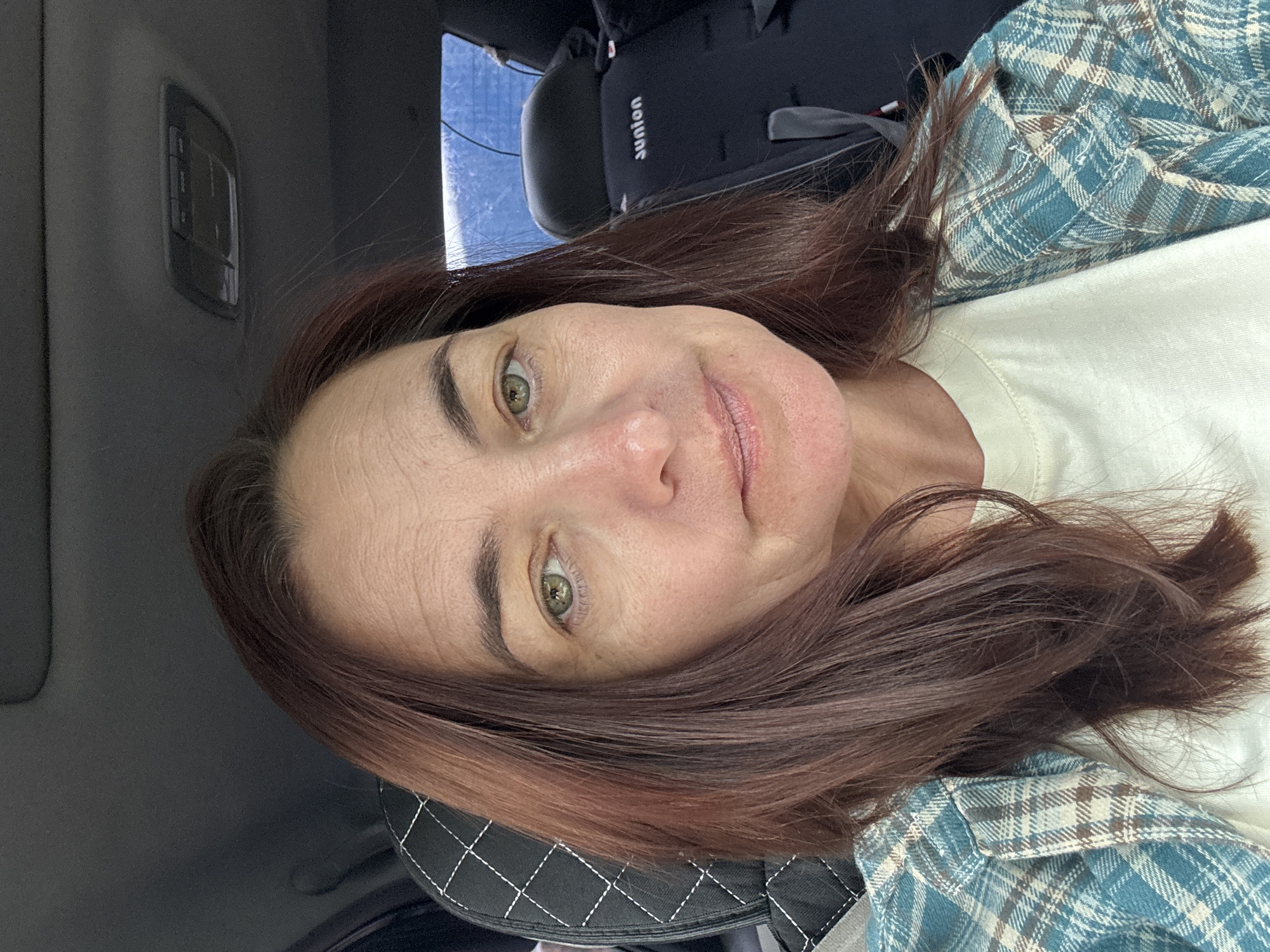 The image is a selfie of a woman inside a vehicle. She has brown hair and green eyes, and is wearing a plaid shirt over a light-colored t-shirt. The background shows car interior elements, including a car seat with the brand "sunion" visible. The image does not contain any information that would allow for the determination of its location in the city of Ufa, Russia.