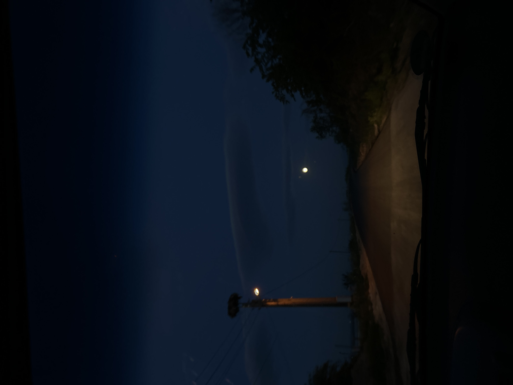 The image depicts a night scene in Ovchepoltsi, Bulgaria. A road stretches into the darkness, illuminated by a distant street light. A utility pole stands to the left, with wires extending toward the sky. In the sky, the moon shines faintly through a thin cloud cover. The surrounding area appears to be rural, with vegetation lining the road. The image seems to have been taken from inside a vehicle.