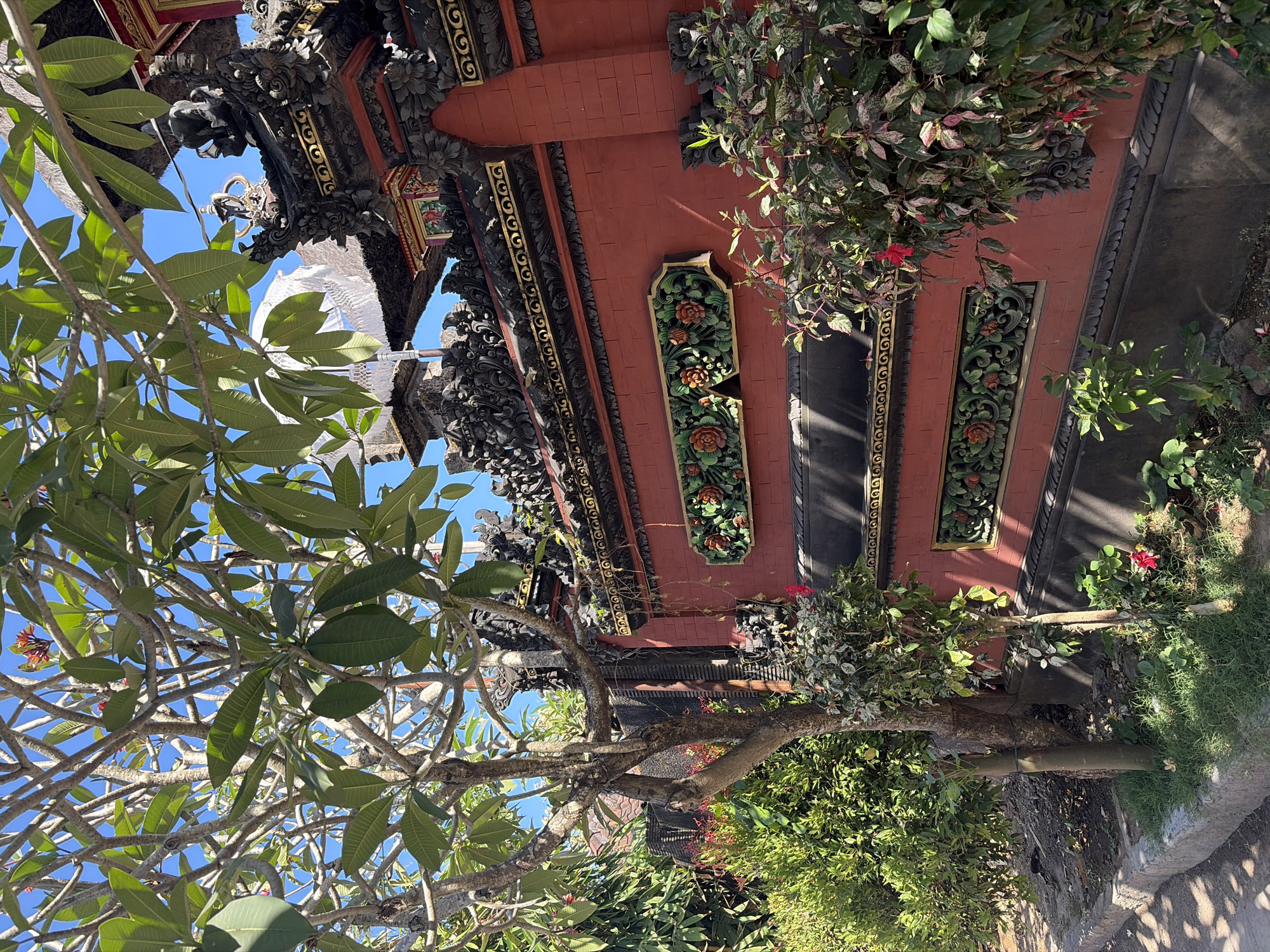 The image depicts an ornate, red brick wall with decorative black and gold trim, characteristic of Balinese architecture, situated in Kuta Selatan, Indonesia. The wall features a carved panel with floral motifs in green and orange, and is topped with intricate dark stone carvings. A lush green tree with broad leaves partially obscures the upper portion of the wall and the sky. To the left of the wall, dense green foliage and a smaller tree with visible branches are present. In the foreground, grass and small plants grow at the base of the wall, with a stone curb lining a pathway. The scene is brightly lit, suggesting a sunny day.