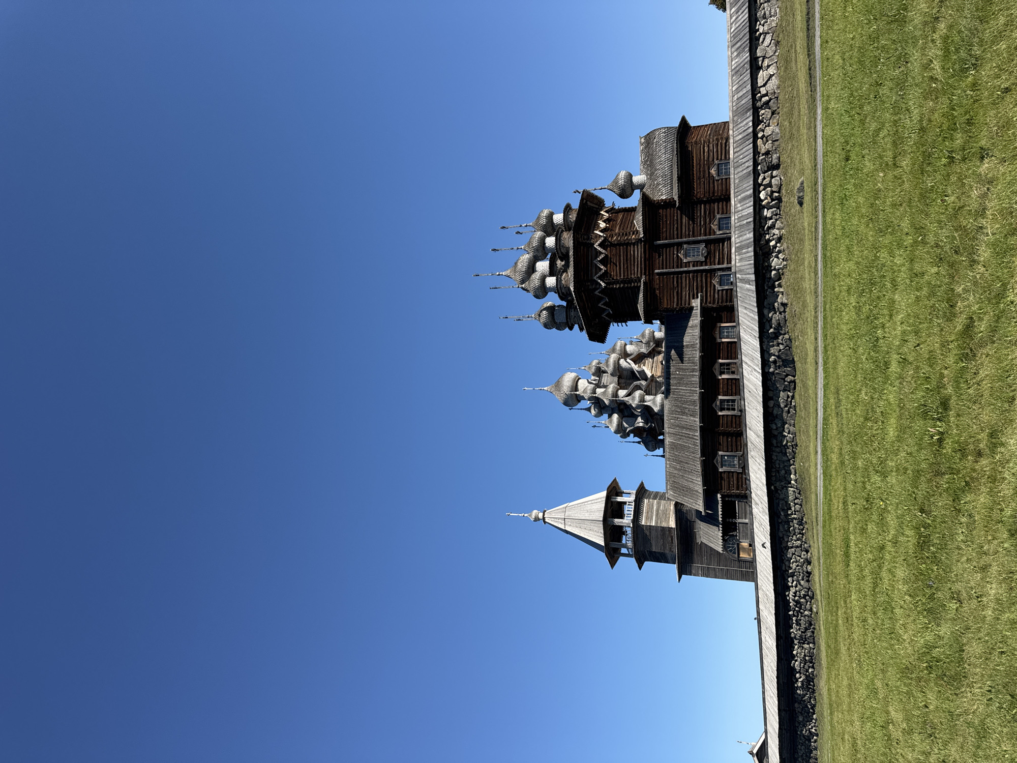 The image captures a low-angle, full-shot view of the Kizhi Pogost in Boyarshchina, Russia. The historic wooden architectural complex, bathed in clear, bright sunlight under a vast, unblemished blue sky, stands prominently. The Kizhi Pogost is characterized by its intricate wooden structures, featuring multiple onion domes and a tall, spire-topped bell tower. A stone wall, topped with wooden fencing, forms a boundary in front of the main structures, separating them from a lush, green grassy field in the foreground. The overall scene conveys a sense of historical significance and natural beauty, with no people or active events depicted.