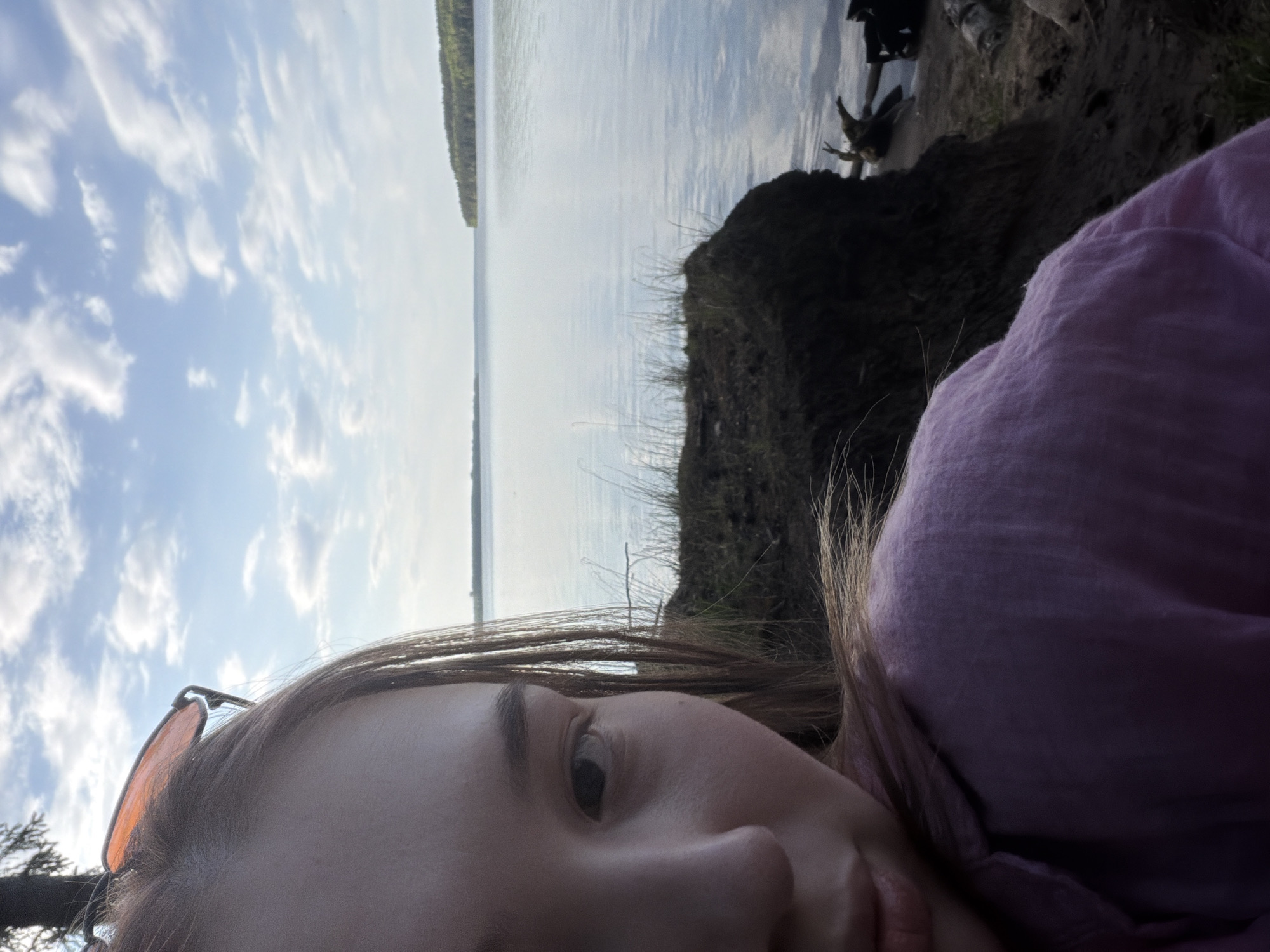 A selfie-style photograph is taken with a low angle, featuring a person lying on what appears to be an embankment or a hillside. The person, likely female, is wearing a lavender-colored garment. Part of her face is visible, including an eye, nose, and mouth, with glasses perched on her head.
Behind the person, the scene opens up to a large body of water, possibly a river or lake, reflecting the sky. On the right, a dark, grassy embankment slopes down to the water's edge. On the opposite side of the water body, a strip of dense forest is visible in the distance. The sky is filled with scattered clouds, suggesting a partly cloudy day.
Location is described as Chaikovsky, Russia. Therefore, the water body may be one of the reservoir in the region.