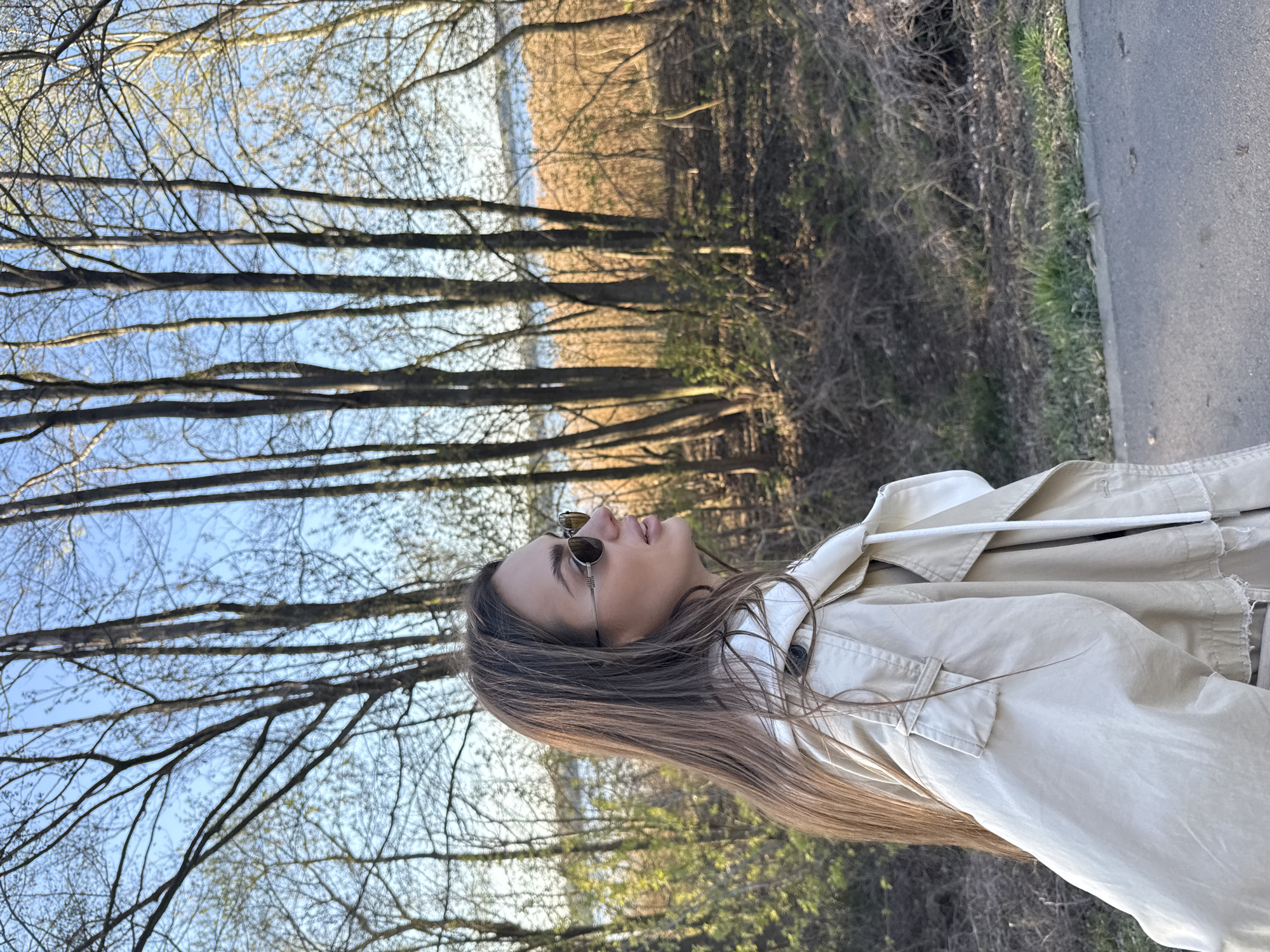 A young woman wearing sunglasses and a beige jacket is the central subject of the image. She is seen in profile, looking upwards. The location appears to be a park or wooded area near a road, with bare trees lining the background. Based on the prompt, this is likely in or near Białobrzegi, Poland. The background includes a body of water visible through the trees. A road with a curb is visible on the right side of the frame.