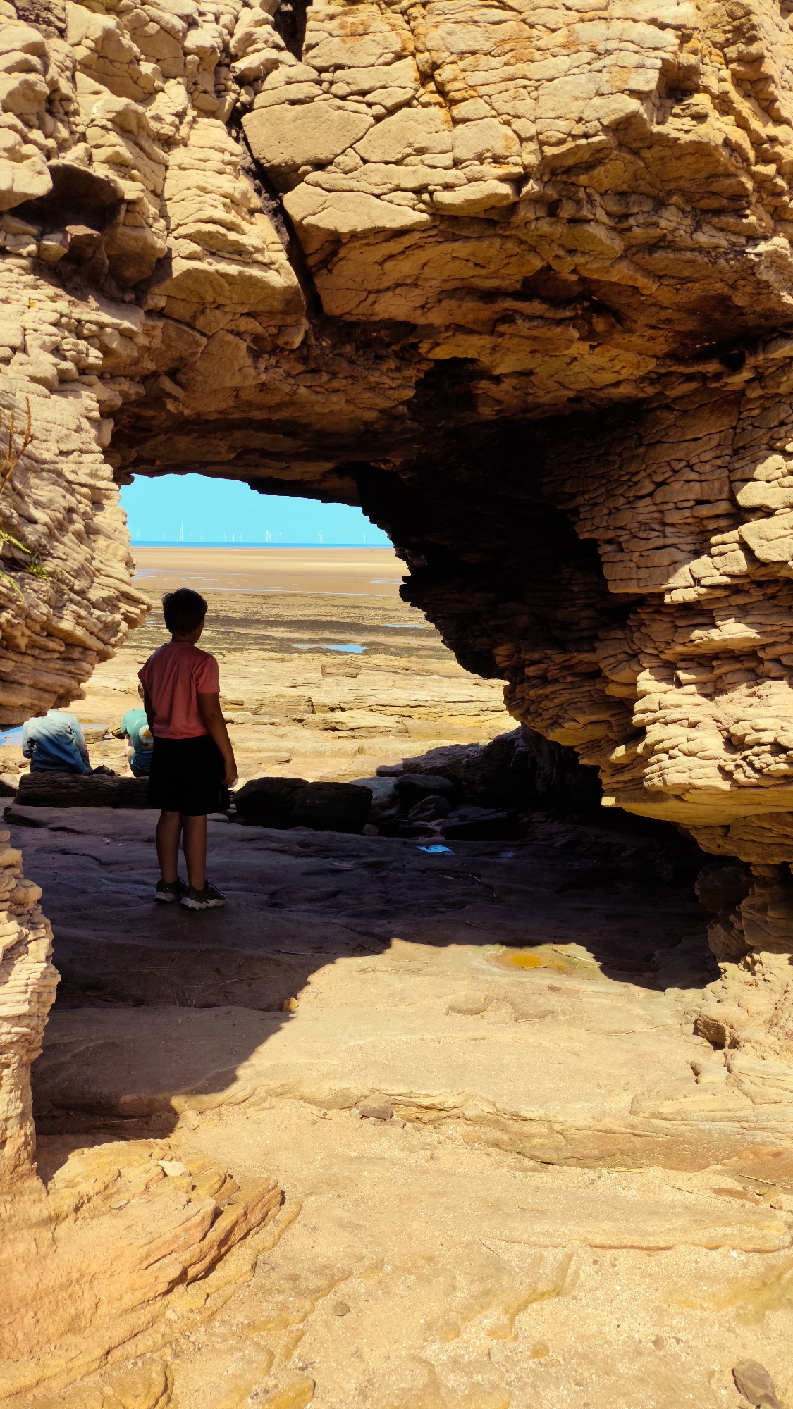 A young person stands in the foreground, facing away from the camera and looking out at a beach scene. They are wearing a pink t-shirt and dark shorts, with dark sneakers on their feet. The person is positioned within a natural rock archway. To the left of the frame, another person is seated, wearing a blue top and dark trousers, with their back to the camera. They are sitting on a dark log. The background reveals a sandy beach with shallow pools of water. In the distance, a line of wind turbines can be seen on the horizon. The rock formations are layered and appear to be sandstone. The overall setting is a coastal environment, likely at low tide. The lighting suggests it is a sunny day. This scene is located at the "Hoylake Docks" area of West Kirby, United Kingdom, a location known for its extensive sand flats and prominent rock formations.