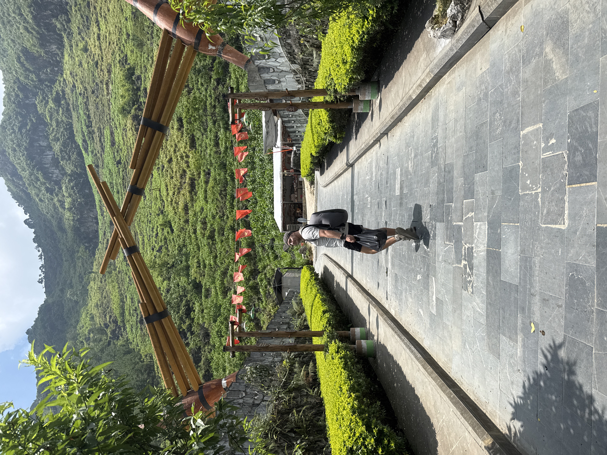 An eye-level shot captured in Mèo Vạc Commune, Vietnam, showcases a paved walkway that curves through lush greenery against a backdrop of a verdant mountainside. A man with a backpack stands on the pathway, his shadow stretching on the gray stone pavement. To the left, two giant crossed wooden instruments, possibly traditional wind instruments, stand upright. A series of wooden posts decorated with small red flags line the path. The walkway is bordered by carefully trimmed shrubs, enhancing the natural aesthetics of the surroundings. The sky is partly visible, suggesting a bright, sunny day.