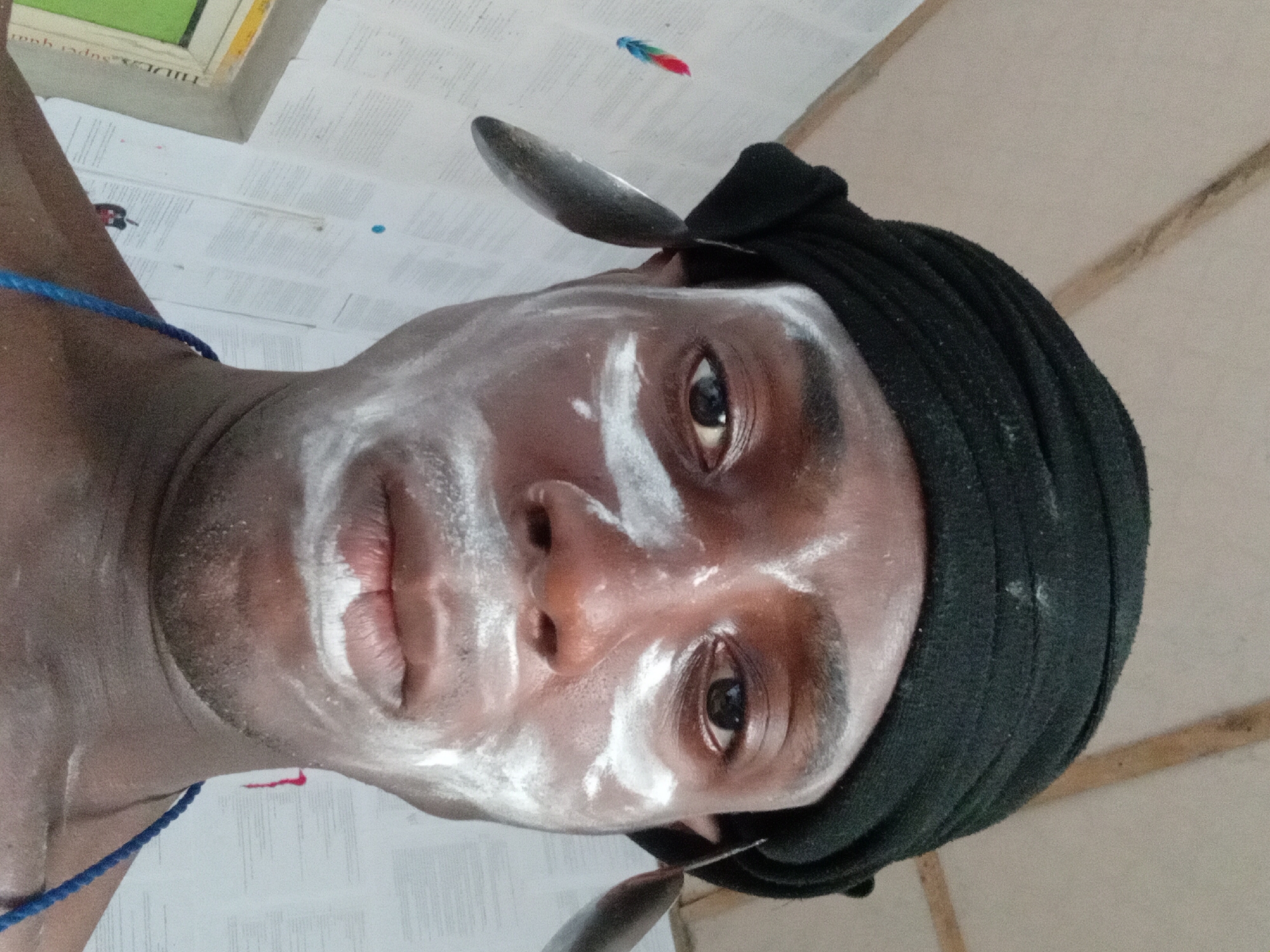 A close-up, slightly angled selfie shot of a man with dark skin, a black head wrap, and white powder-like substance smeared on his face. He has a spoon balanced on each side of his head, appearing to be held in place by the head wrap. A blue rope is visible around his neck. The background consists of papers on a ceiling, with a window frame in the upper left and a corner of a wooden structure on the right. The image appears to be taken indoors in an area with poor lighting. The location is unknown.