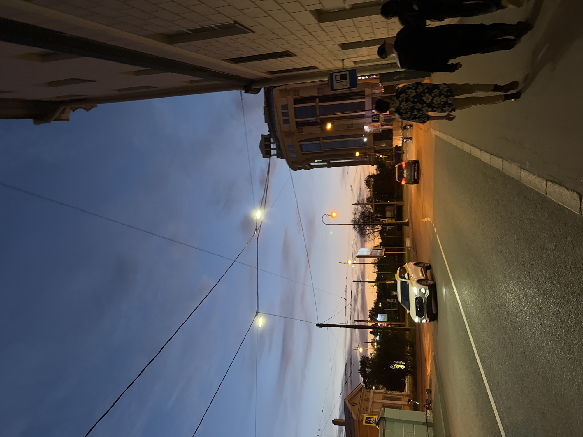 Here's a description of the image:
The image depicts a street scene in Saint Petersburg, Russia at dusk. The sky transitions from a deep blue to a pale orange near the horizon. Overhead power lines stretch across the sky. On the right side of the frame, there is a paved road with a white line marking the lane. A white car is driving on the road. Further down the street, there's a group of people walking on the sidewalk, and cars further away. Buildings line both sides of the street, one a classic, ornate building, the other with a more modern look with a bus sign. Streetlights illuminate the scene. A taxi stand can be seen.