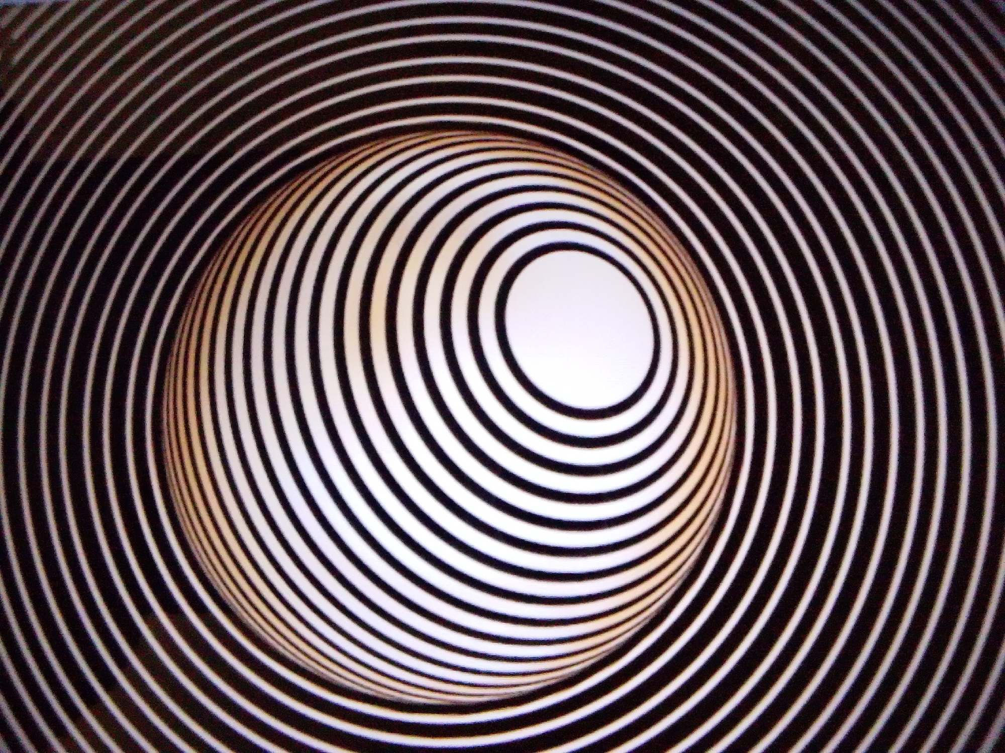 This is an abstract, optical illusion image featuring concentric circles that create a sense of depth and a warped spherical shape. The circles alternate between black and white, with a yellowish tint visible on the curved surface. The overall effect is that of a three-dimensional tunnel or funnel. There are no people, identifiable objects, or environmental features in the image. The location is unknown, and no city or street is mentioned.