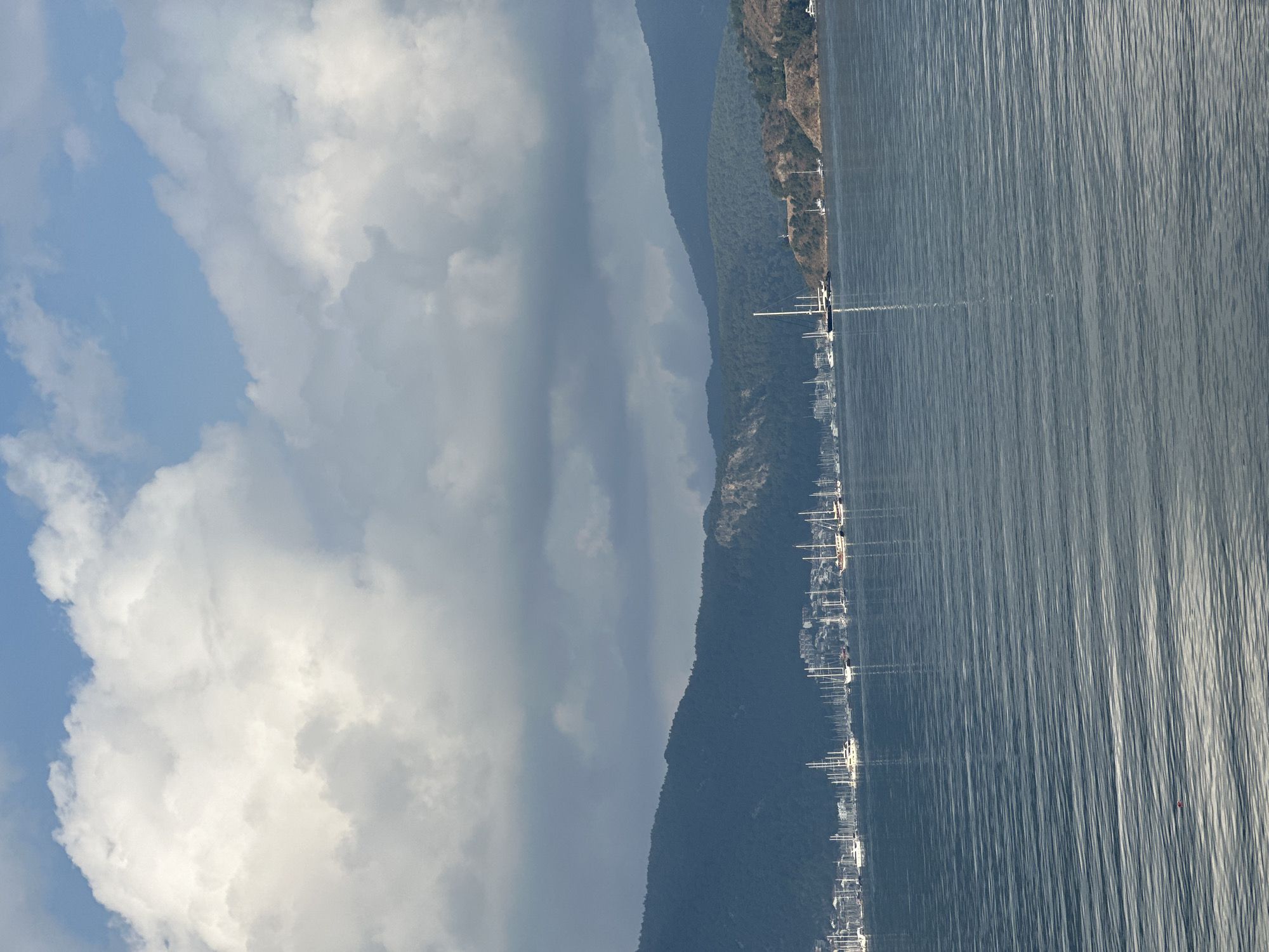 The image shows a view of Fethiye, Turkey, likely from a high vantage point. The foreground is dominated by the sea, its surface rippled by the wind. Moored sailboats line the coastline, their masts creating a vertical pattern against the backdrop of the sea and the adjacent land. Lush, green hills rise along the coast, forming a natural barrier between the town and the open water. The sky is partly cloudy, with large, billowing clouds contrasting against patches of blue sky. The overall impression is one of a scenic, coastal landscape with a marina or harbor.
