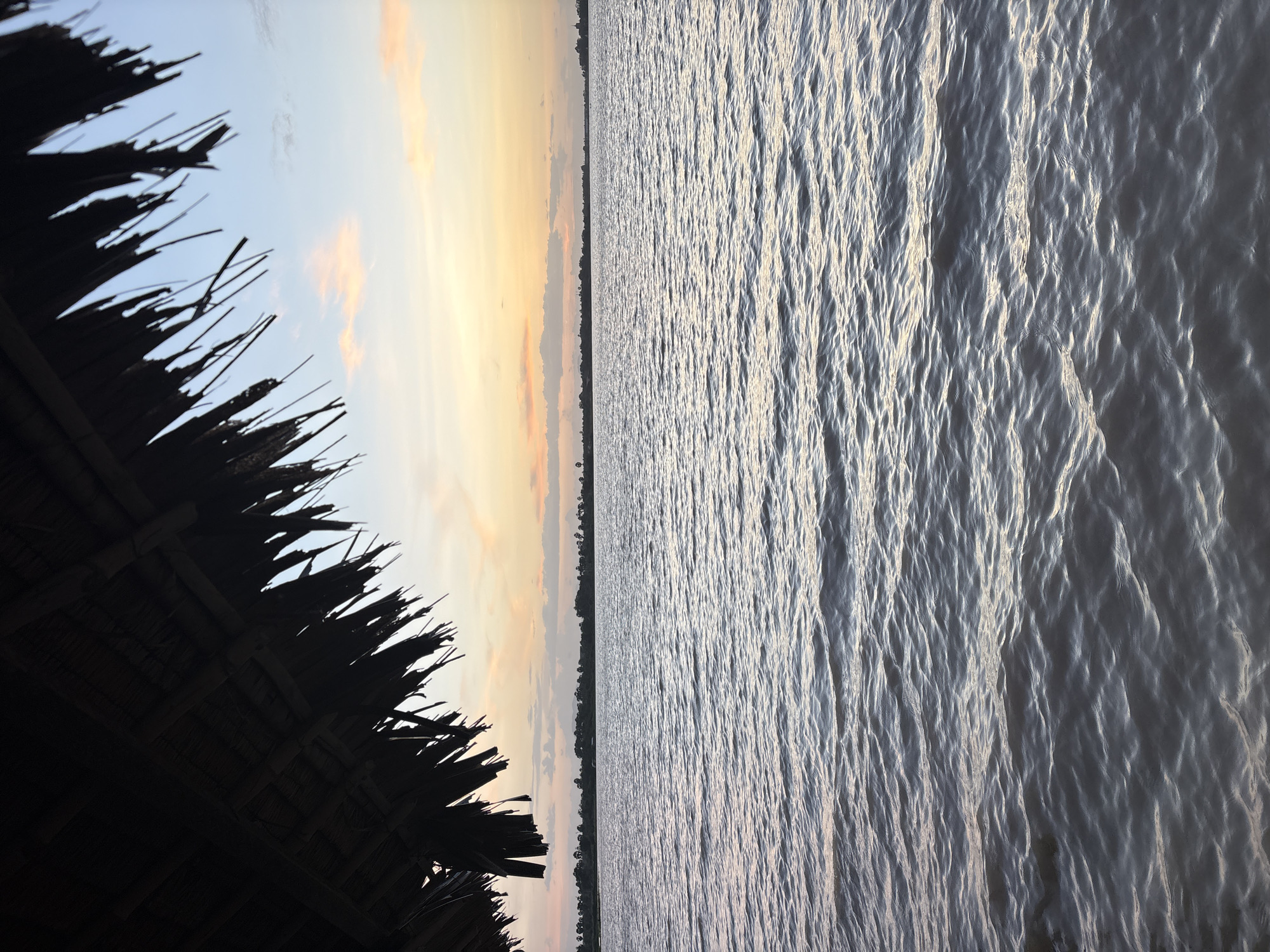 The image shows a body of water, likely a river or lake, with gentle ripples. In the foreground, the thatched roof of a shelter juts in from the upper left corner, casting a shadow. Beyond the water, a tree-lined horizon is visible under a sky with soft orange and pink clouds, indicative of a sunset or sunrise. The location is specified as Chhômprâk Trâb, Cambodia. There are no people or animals visible in the image. The environment is natural, with the focus on the water, sky, and the rustic shelter.