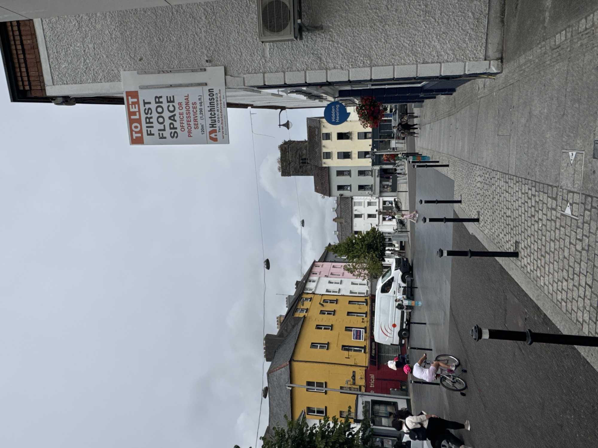 The image depicts a street scene in Waterford, Ireland, on a cloudy day. On the right side of the image, a textured wall of a building is visible, with an air conditioning unit mounted high on it. A white sign with red text is attached to this wall, advertising "TO LET FIRST FLOOR SPACE OFFICE OR PROFESSIONAL SERVICES". Below this, the name "Hutchinson" appears, along with a phone number and website address.
To the left of the wall, a sidewalk is visible, paved with large grey flagstones and bordered by cobblestones. A dark blue building runs along the sidewalk, with a circular blue sign that reads "Hidden Hearing". Hanging baskets with red flowers are placed near the "Hidden Hearing" sign. Further down the sidewalk, people are walking.
Across the street, a row of buildings with colorful facades is present. The building closest to the foreground on the left is yellow with dark shutters and red signage. Further down the street, buildings in white, pink, and grey are visible. A white van is parked in the street. A young person wearing a pink helmet is riding a bicycle with training wheels on the street. Other pedestrians are also present on the street and sidewalk. The street is paved with asphalt and cobblestones. Lampposts are visible, and overhead wires are strung across the street. In the background, a tall stone tower, likely part of a church, is visible above the buildings.
