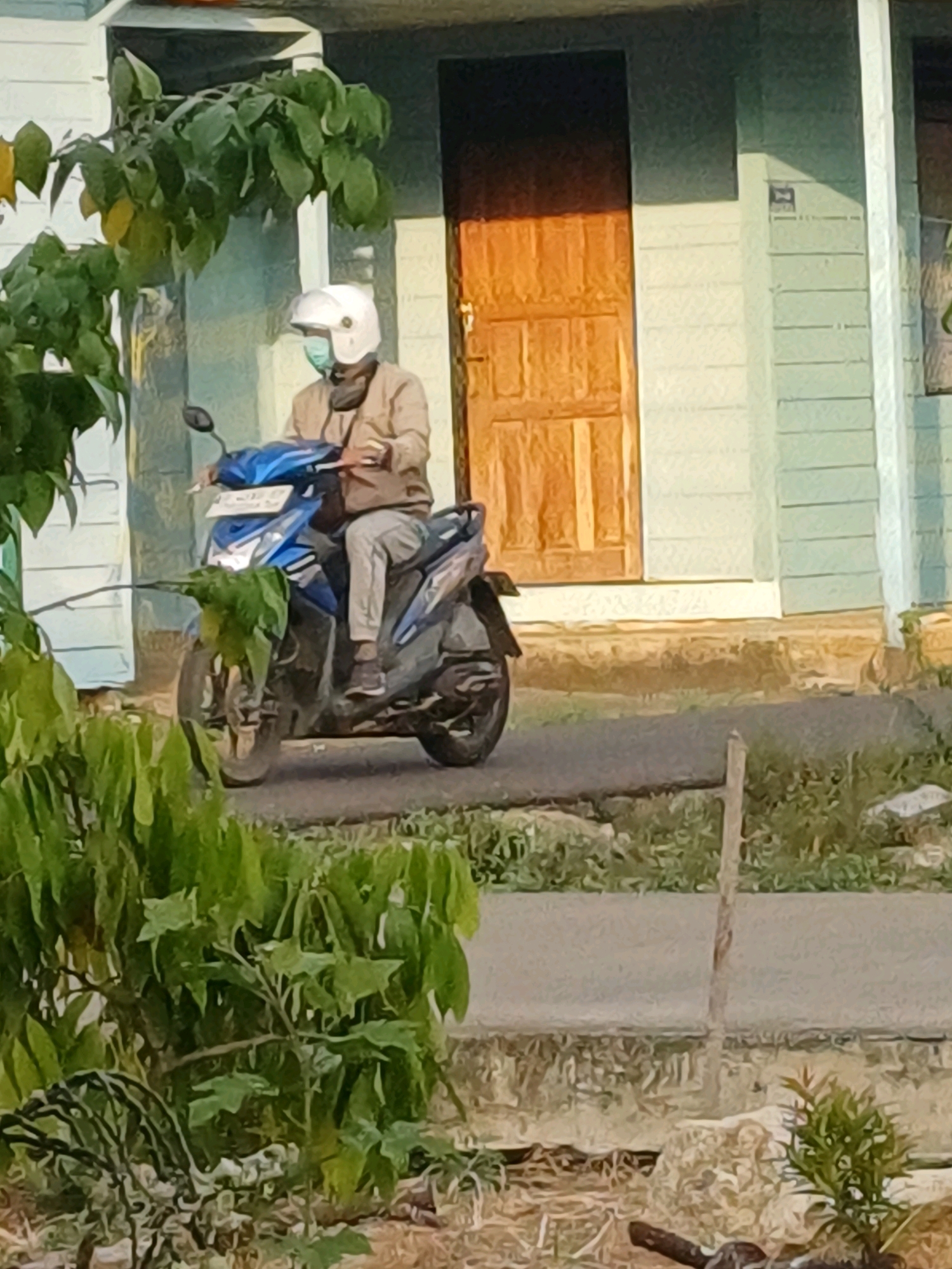 A person wearing a white helmet and a light brown jacket is riding a blue and black scooter. The person is also wearing a light blue mask. The scooter is parked in front of a light-colored building with a wooden door. The building has light green siding above a light-colored base. The scooter has a license plate that is not legible. There is foliage in the foreground and to the left of the scene. The ground in front of the building appears to be paved. The background shows a wall and a door, with greenery on the left side. The image is taken from a slightly elevated perspective, looking down at the scooter and rider. The overall scene suggests a residential or commercial area in Pasir Belengkong, Indonesia.