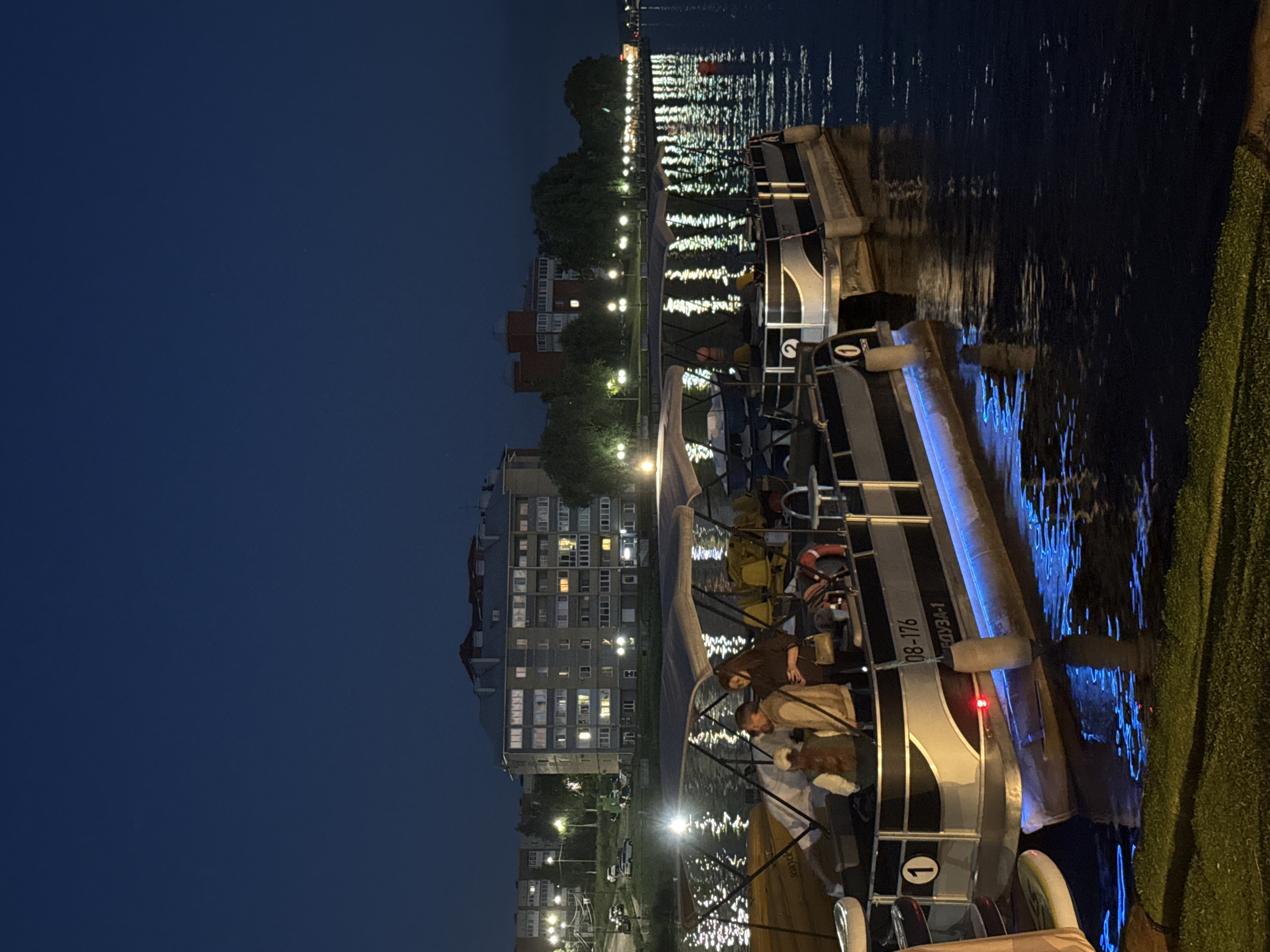 At night, several pontoon boats are docked on the dark water of a lake in Votkinsk, Russia. The boats are equipped with canopies and some have illuminated blue lights under their hulls, reflecting on the water. People are visible on the boats, some standing and conversing, while others appear to be seated. In the background, a multi-story building with illuminated windows is visible, along with streetlights and trees lining the shore. The sky is dark, suggesting it is nighttime. The scene appears to be a waterfront area or marina in the city.