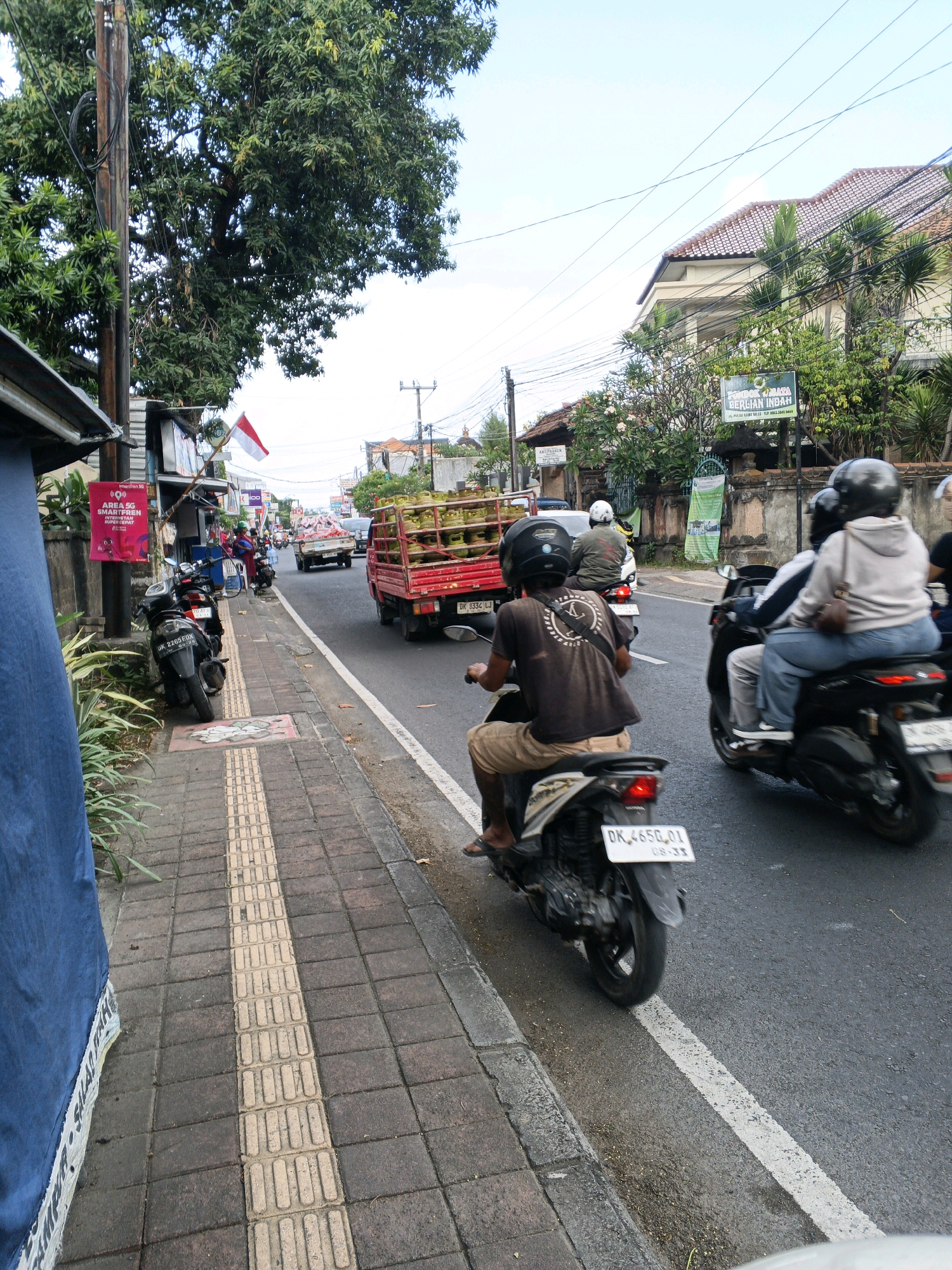 The image depicts a street scene in Denpasar, Indonesia, likely on a main road with moderate traffic. In the foreground, a person rides a motorcycle, their back to the camera, wearing a brown t-shirt and light-colored shorts. The motorcycle has a license plate that reads "DK 4650 QJ". To the right, two other individuals are on a motorcycle, with the passenger in a light-colored hooded sweatshirt and jeans. In the background, a red truck is transporting a load of green produce, possibly coconuts, with a license plate visible as "DK 3332 LL". Other vehicles, including cars and motorcycles, are also present on the road, suggesting ongoing activity. The street is lined with buildings on the right, featuring tiled roofs and lush vegetation. A sidewalk runs along the left side of the street, paved with interlocking bricks and a tactile paving strip. A utility pole with wires runs alongside the sidewalk, with an Indonesian flag displayed. The overall impression is a typical urban street environment in Denpasar.