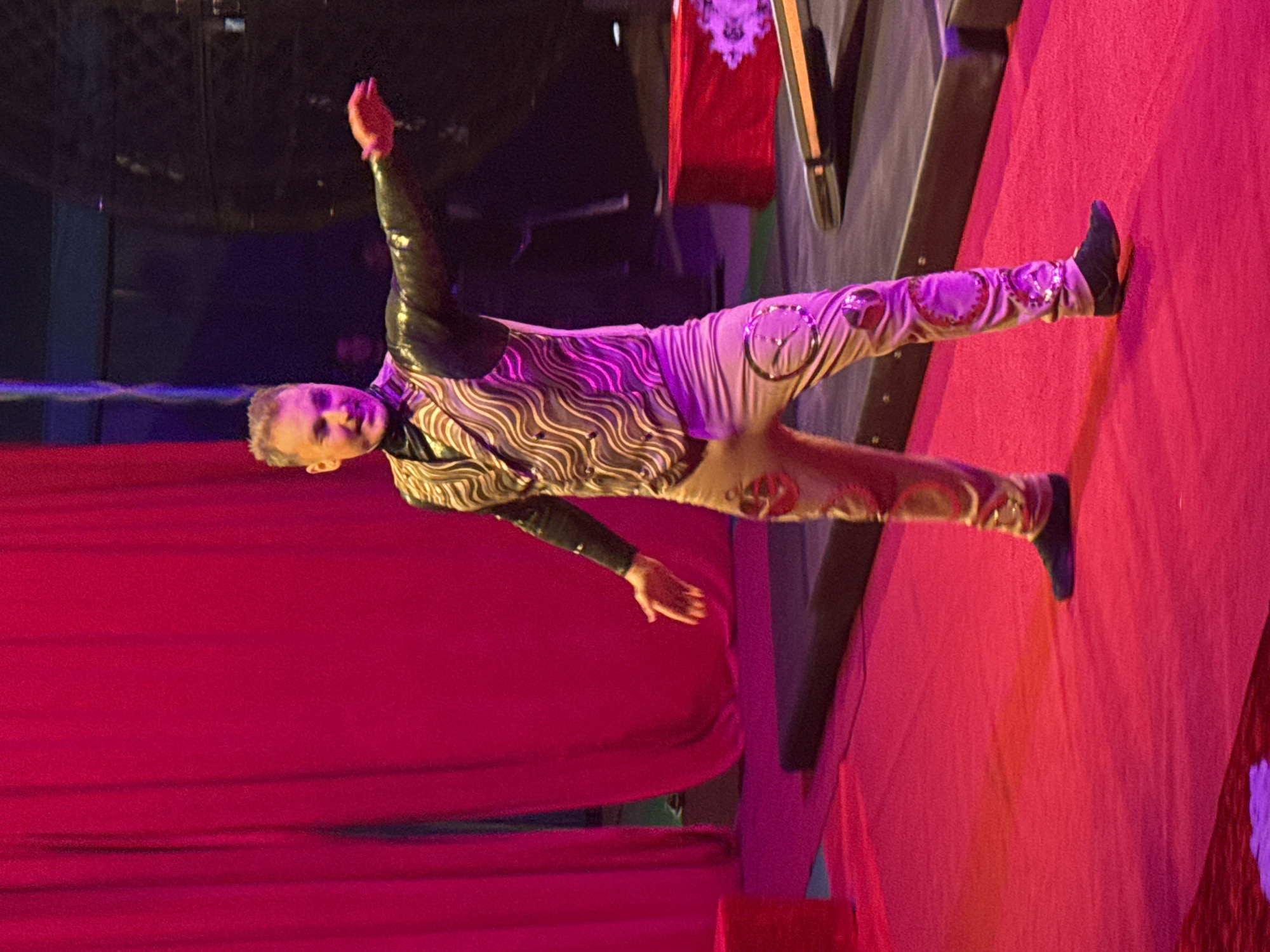 The image depicts a male performer, possibly an acrobat, balancing horizontally with one foot placed on a red surface. He is wearing a costume with brown and silver wavy patterns on the torso and cream-colored pants adorned with circular designs. The background includes a red curtain, a black structure, and a red and white banner. The lighting is theatrical, with red and purple hues. The location is most likely a circus or theater stage and is not possible to place in the city of Dmitrov, Russia, without further information.