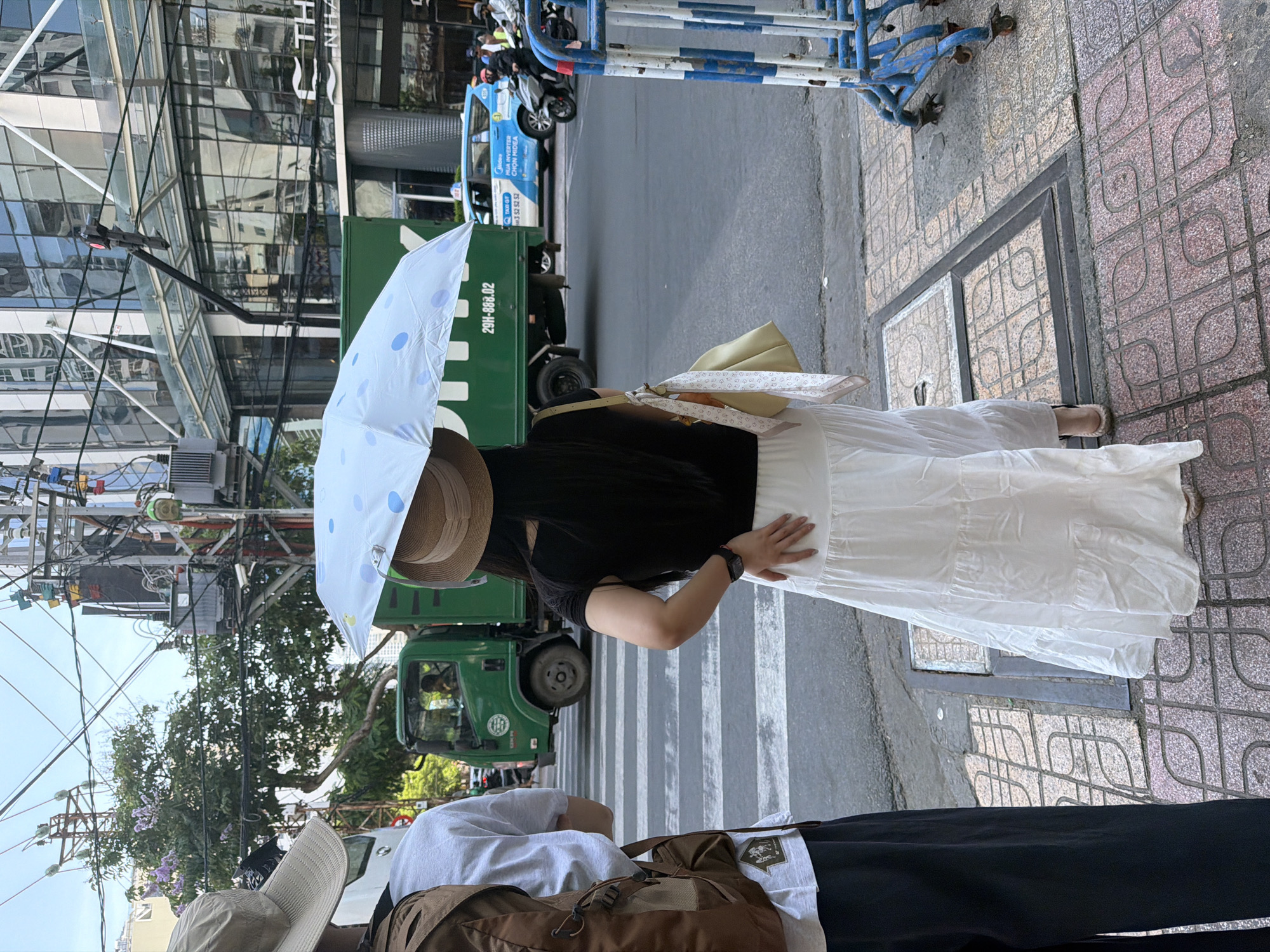 A woman wearing a hat and a white tiered skirt stands on a sidewalk in Nha Trang Ward, Vietnam, holding a white umbrella with blue polka dots. She is facing away from the camera, her right arm bent at the elbow. A person wearing a beige hat and a backpack stands to her left, also facing away from the camera. A green truck is stopped at a crosswalk behind them. A blue taxi is visible in the background, along with several motorcycles and a modern building. A section of blue and white barricade is visible on the right side of the image. The ground is paved with decorative tiles, and there are several manhole covers. The sky is overcast.
