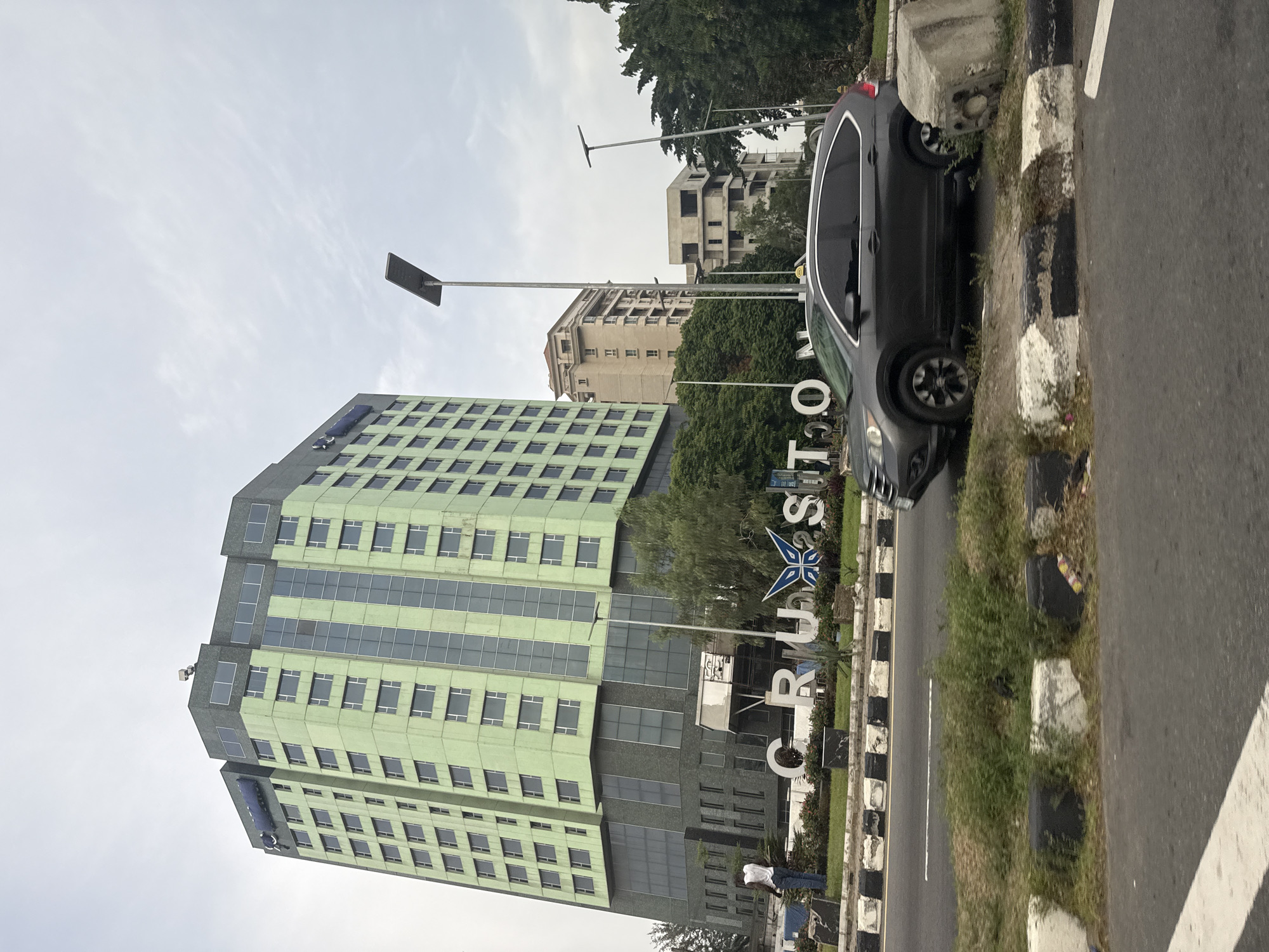 The image captures a daytime scene in Falomo, Nigeria. A tall, green and grey striped building dominates the left side of the frame. In front of the building is the white 'CRUSTON' signage. To the right of the signage, on the side of the road, is a black car parked at an angle, with its tires pressed against the curb. The road has a black and white patterned curb and a painted white line. Other buildings can be seen in the background. A dark grey sky is visible overhead. A light pole is present. A person is standing near the 'CRUSTON' signage.