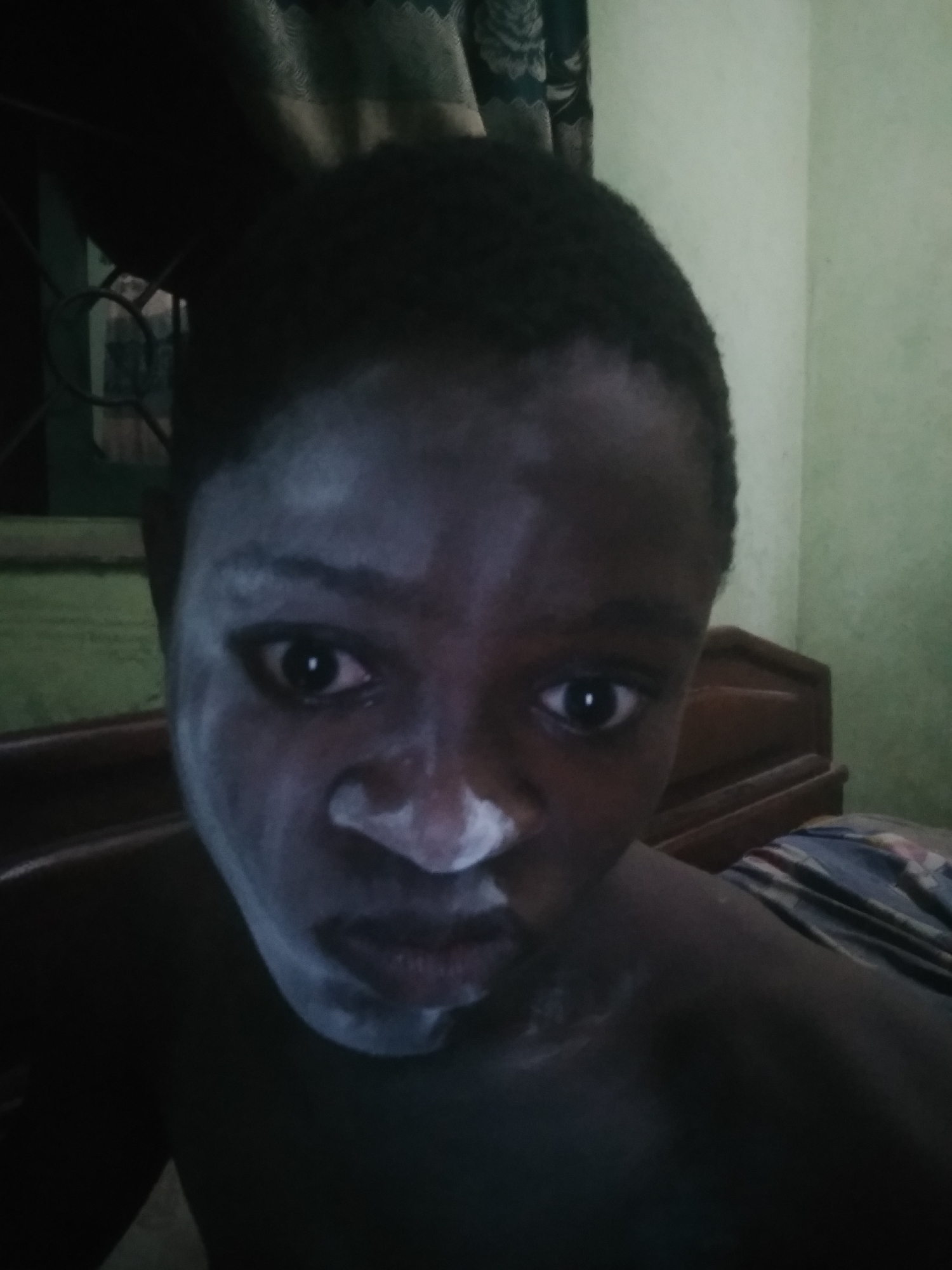The image is a close-up of a person's face, partially covered in a white substance resembling powder. The person has dark skin and short, dark hair. Their eyes are wide and appear to be looking directly at the camera. The white substance is concentrated around the forehead, nose, and mouth. The background is blurry but suggests an interior setting. A wooden headboard of a bed is visible behind the person, and a window with a curtain can be seen in the upper left corner. The wall appears to be light green. There is no immediate indication of the location being Shangisha, Nigeria within the visual elements of the image.
