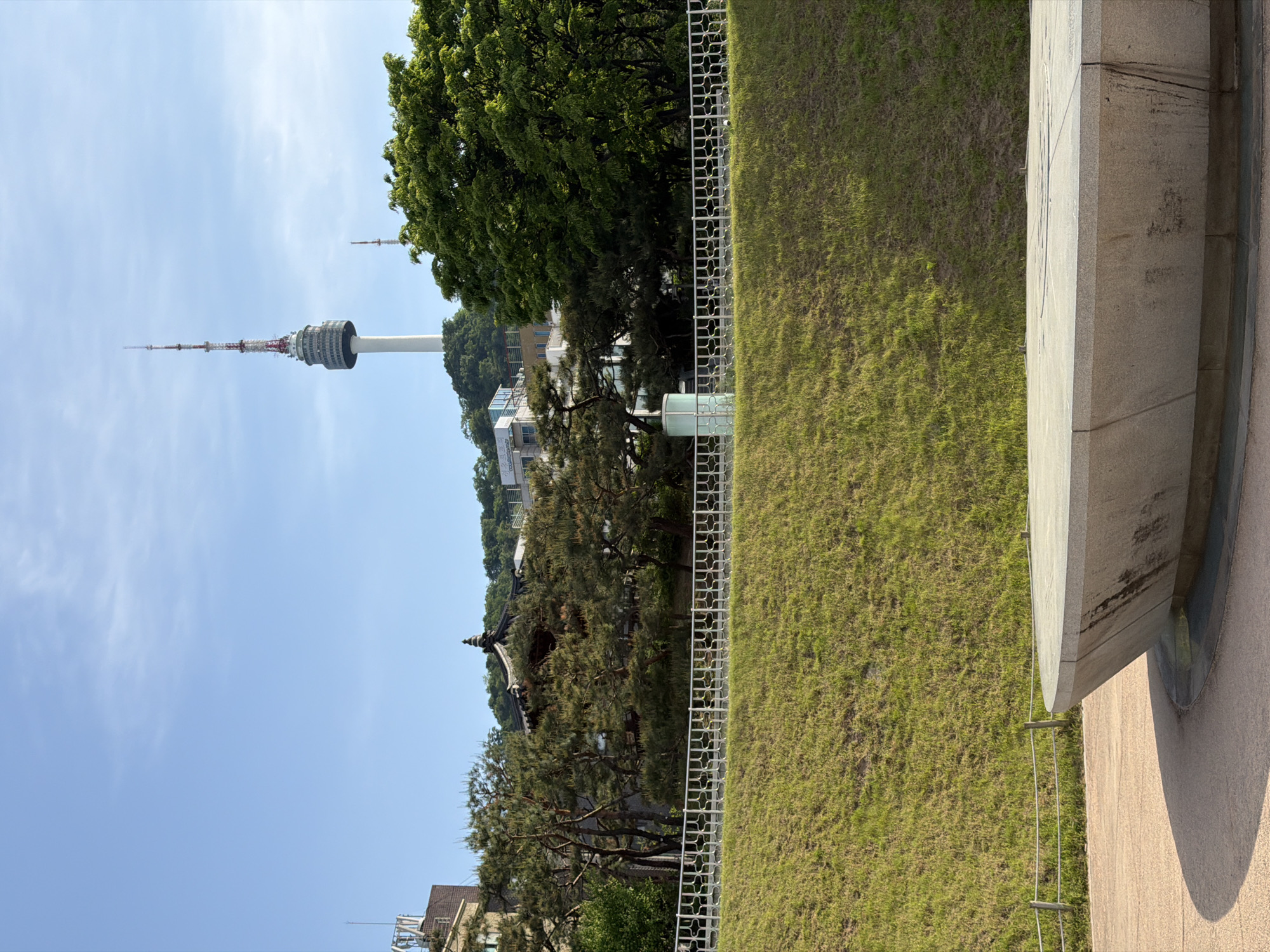 This outdoor shot, taken during daylight in Seoul, South Korea, showcases the N Seoul Tower in the distance. To the right, a stone structure and grassy slope are visible, with a white fence separating the grass from trees and buildings. The sky is a clear blue with light wisps of clouds. The tower is a focal point, positioned against the sky with its recognizable structure.
