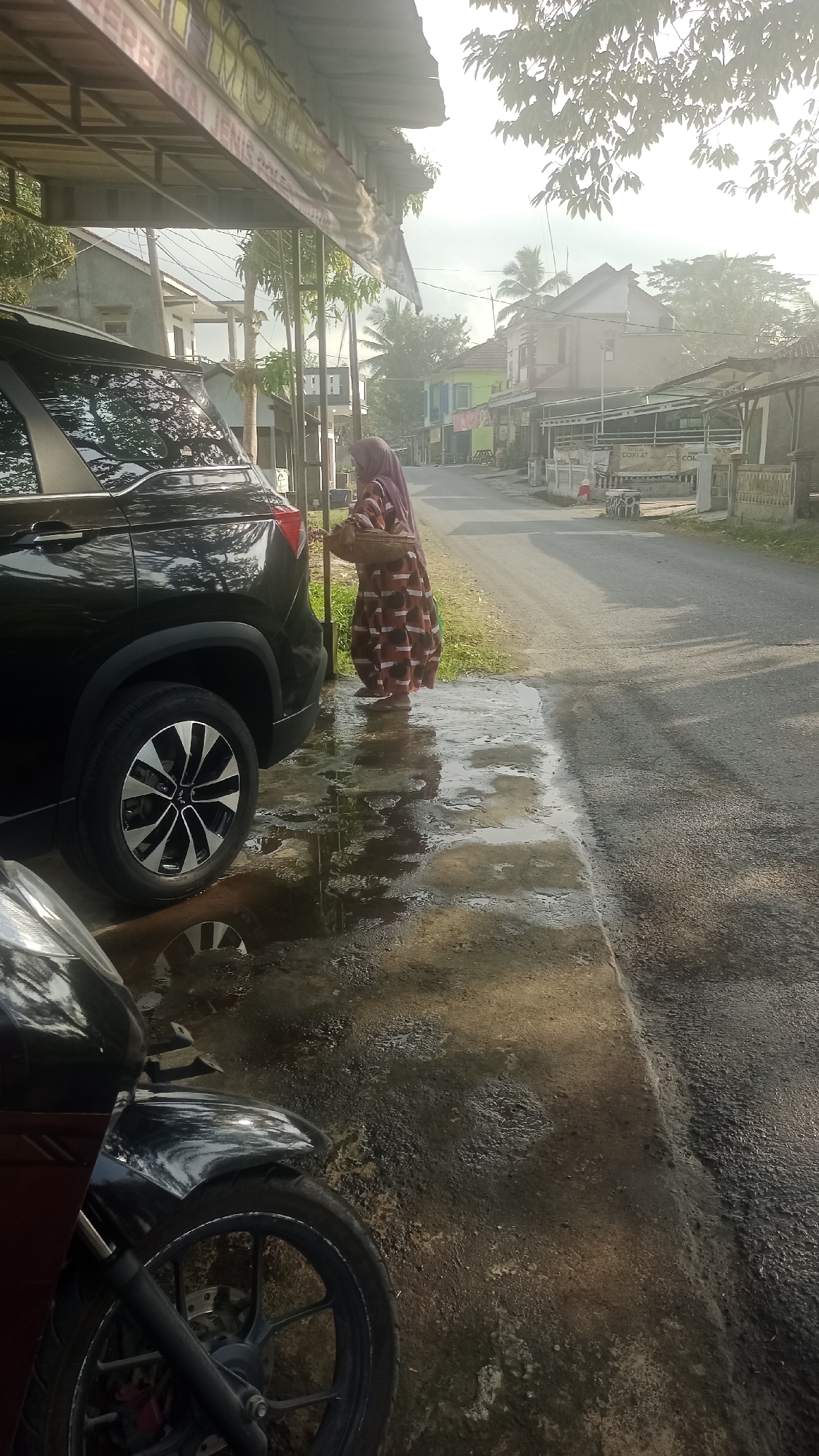 A woman wearing a headscarf and a patterned dress stands on the side of a road in Tonjong, Indonesia. She is holding a woven basket and appears to be interacting with a black SUV parked nearby. The ground is wet, with puddles reflecting the sky and surrounding environment. A motorcycle is partially visible in the foreground, also on the wet ground. The road is lined with buildings and trees, suggesting a residential or commercial area. The lighting indicates it might be early morning or late afternoon.