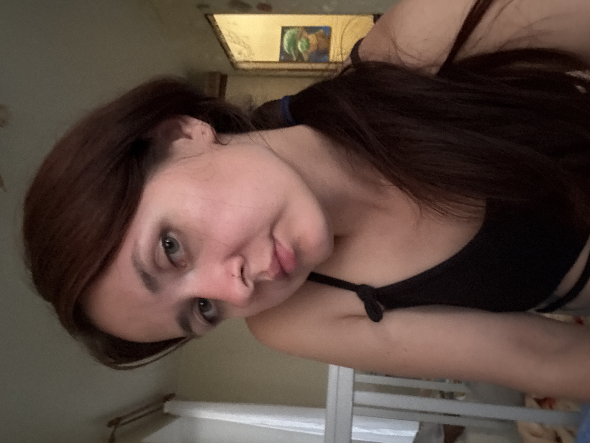 A woman with dark brown hair tied in a ponytail and light green eyes is looking towards the camera. She is wearing a black tank top. Her left shoulder is visible, and she has a nose piercing. In the background, there is a crib with white railings. To the right, a doorway is visible, with a framed picture of a character that appears to be from a popular culture franchise hanging on the wall. The overall lighting suggests an indoor environment, possibly a bedroom. The image does not provide specific geographical markers to confirm a location in Naberezhnye Chelny, Russia.