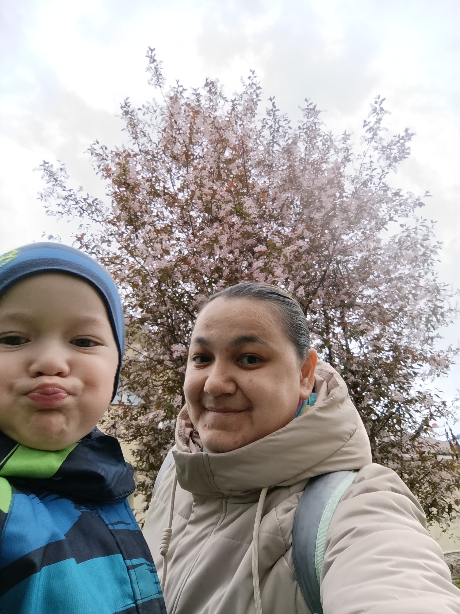 A medium shot captures a woman and a child, likely a selfie, taken outdoors on an overcast day. The child, wearing a blue and black striped jacket and a blue hat, is on the left with pursed lips. To the right, a woman in a beige jacket smiles slightly. Behind them is a tree in bloom with small, light pink blossoms against a cloudy, overcast sky. The location is identified as Chaikovsky, Russia.