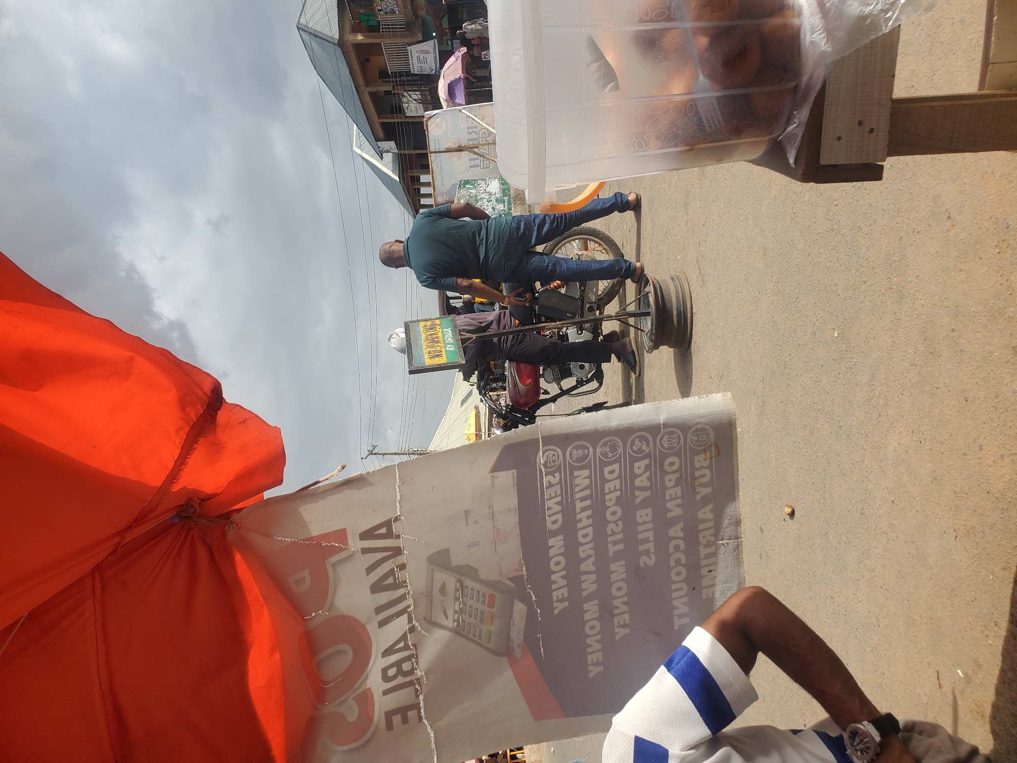 In Gwong, Nigeria, a street scene unfolds under a cloudy sky. An orange canopy provides shade for a sign advertising services such as "SEND MONEY," "WITHDRAW MONEY," "DEPOSIT MONEY," "PAY BILLS," and "BUY AIRTIME." A man in a dark green shirt and blue jeans stands near a motorcycle, facing away from the camera. Another person, partially visible in the foreground, wears a white and blue striped shirt and a watch. In the background, buildings with corrugated metal roofs line the street, and a plastic container filled with food items sits on a wooden surface. The ground is paved with a light-colored, sandy material.
