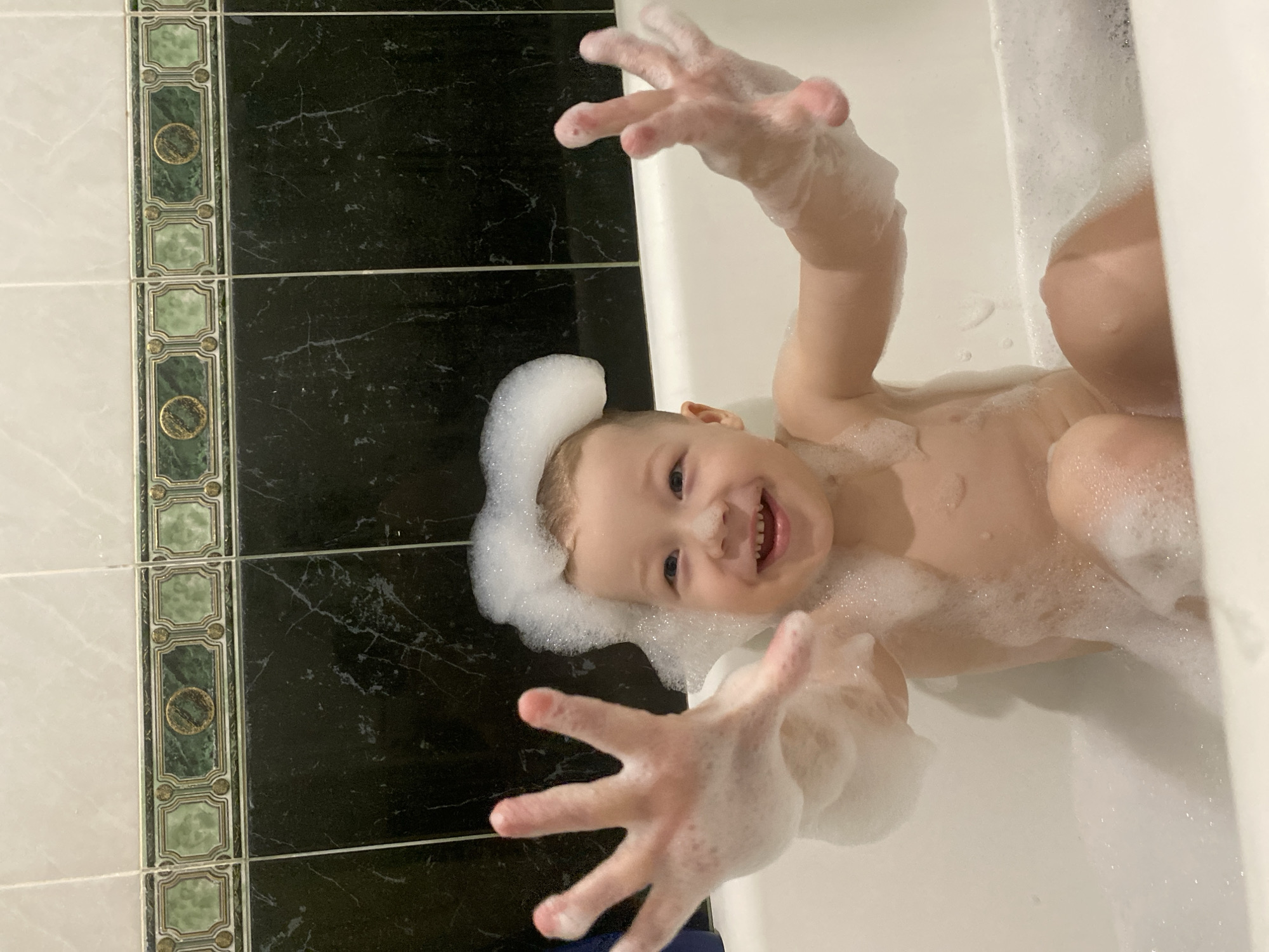A young child, covered in soap suds, is lying in a bathtub. The child is smiling and reaching toward the camera with both hands. Some of the soap suds are piled on the child's head. The bathroom has black tiles with white marbling. A decorative border of green tiles with gold embellishments runs along the edge of the black tiles. The location of the image is Sochi, Russia.