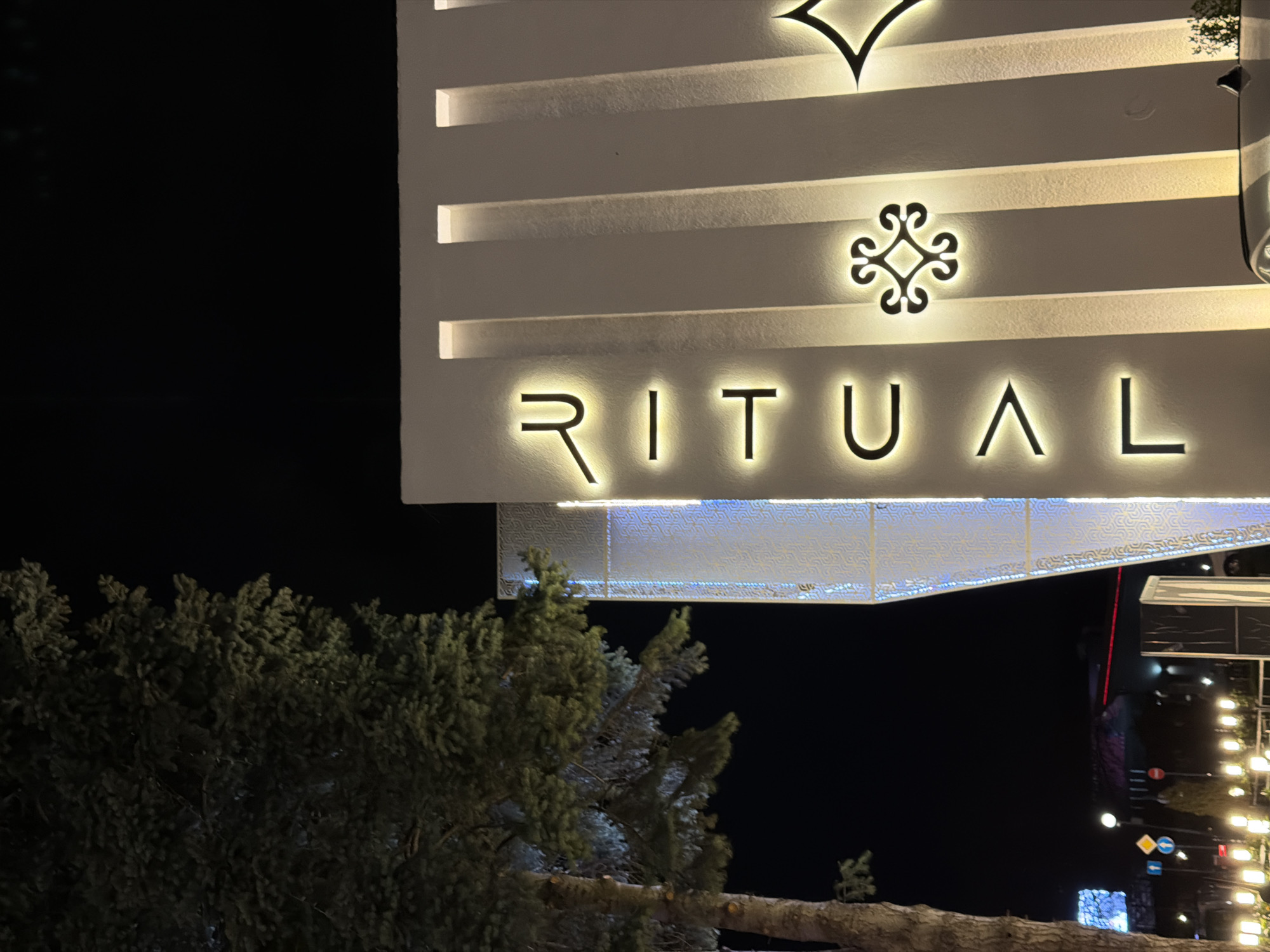 The image shows the "Ritual" building at night in Sofia, Bulgaria. The name "RITUAL" is illuminated in glowing letters on the building's facade. Above the name are two decorative symbols: a stylized flower-like design and a downward-pointing V-shape. The building's design includes horizontal lines with embedded lights. A tree is visible in the lower left foreground. In the background, blurred lights and the silhouette of other buildings suggest a street scene.