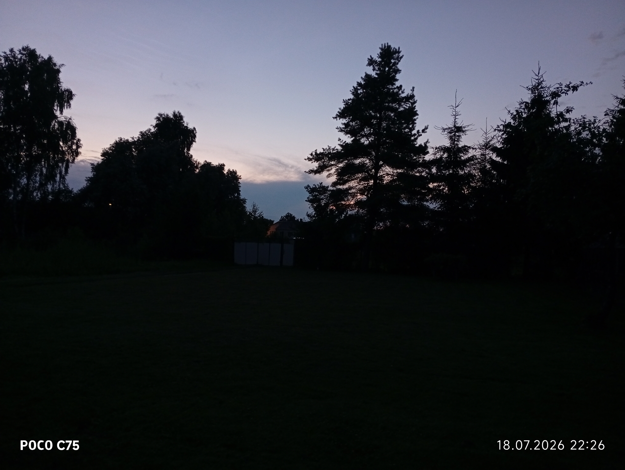 This is a night shot of a grassy field with silhouetted trees and a building in the background. The sky is a deep blue fading to a lighter blue at the horizon, with some faint clouds. The building appears to be a storage unit or shed. The image includes a timestamp and model name in the bottom left and right corners. The location is not specified to be in Daugavpils, Latvia.