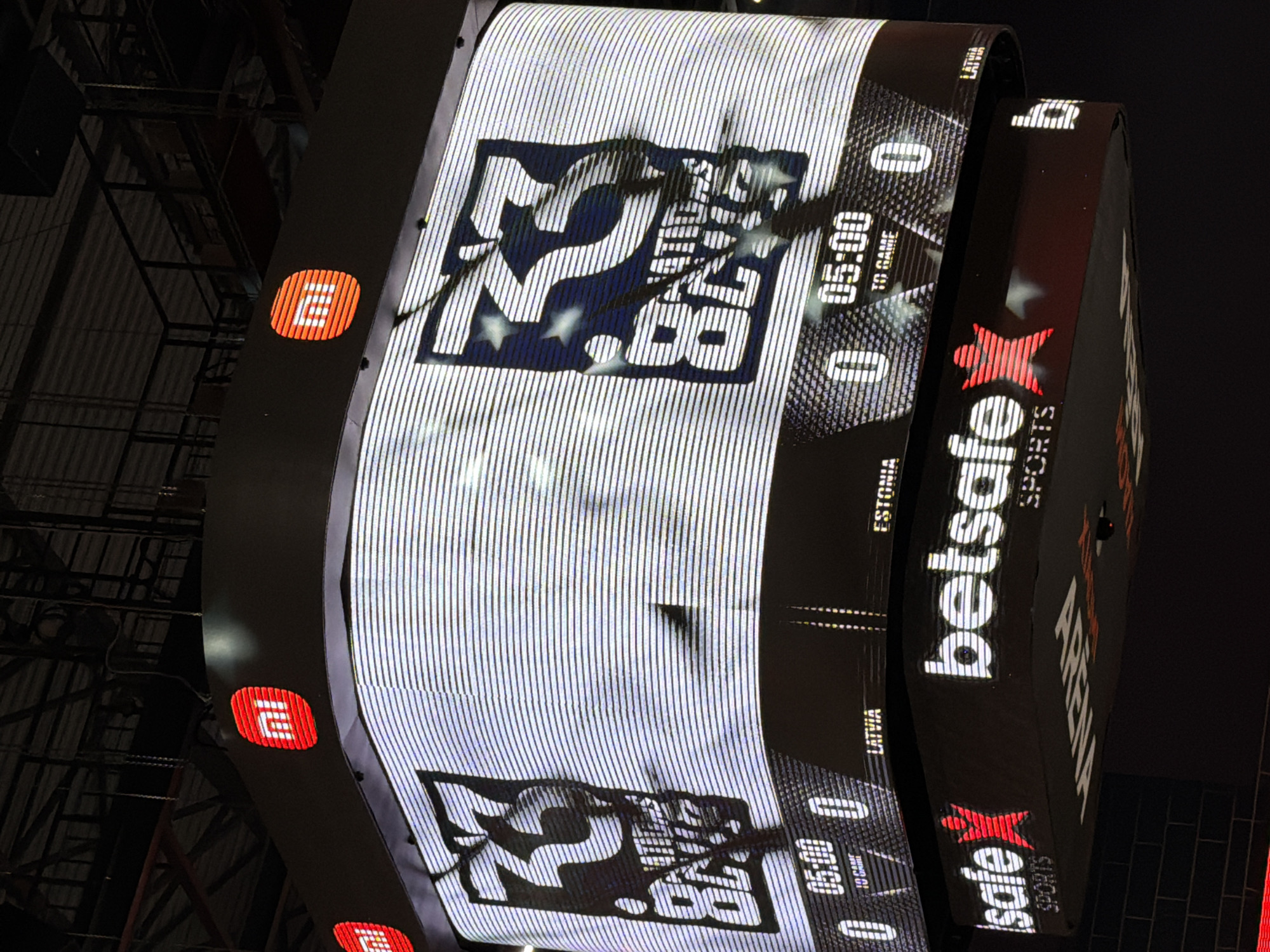 The image shows a close-up of a large, illuminated scoreboard in an indoor sports arena. The scoreboard displays a logo with the number "33" and text that reads "NATIONS LEAGUE". Below this, it shows game information with scores and times, including "05:00" and country names like "LATVIA" and "ESTONIA". There are also logos for "mi" and "betsafe SPORTS" visible on the structure. The overall environment appears to be a modern arena with hanging infrastructure and dark, shadowy areas in the background, suggesting an event is either in progress or about to begin. The presence of Latvian and Estonian team indicators implies the event is related to a sporting competition between these nations.