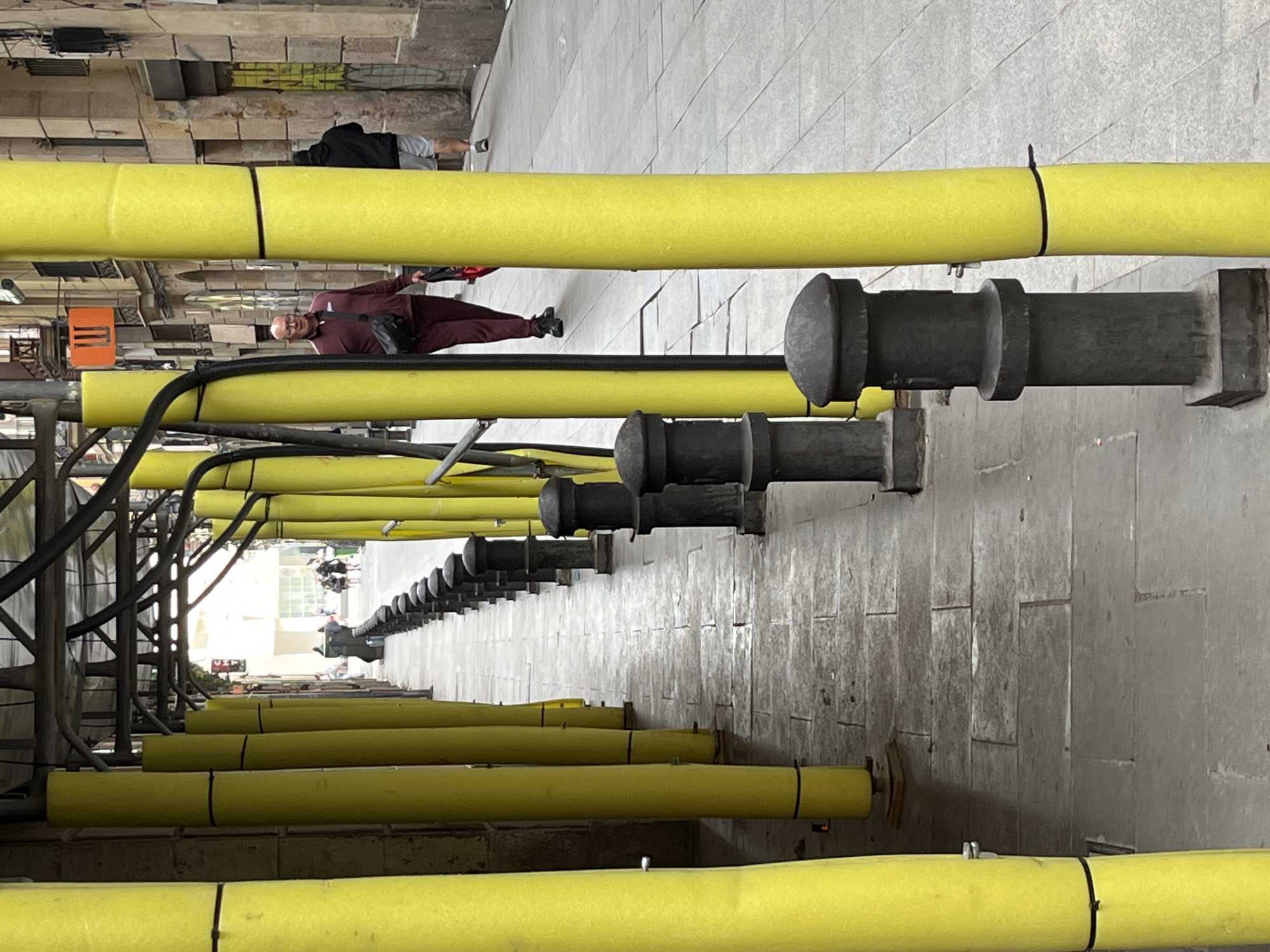 This image captures a long, narrow urban passage, likely an alleyway or a covered walkway, in Barcelona, Spain. The scene is dominated by multiple bright yellow pipes, which appear to be part of an exposed utility system or construction scaffolding. These pipes run horizontally through the frame at various depths, creating strong leading lines that converge towards a distant, brightly lit opening at the far end of the passage.
On the right side, numerous black, vertical pipe-like structures, each topped with a rounded, decorative cap, are affixed to a textured grey wall. These structures also extend into the distance, parallel to the yellow pipes. The left side of the passage features a more complex arrangement of scaffolding-like supports and black cables. The ground is paved with light-colored tiles or concrete.
Two individuals are clearly visible in the mid-ground. Closer to the foreground, a man wearing a maroon (or burgundy) tracksuit, glasses, and a dark shoulder bag is seen walking towards the viewer. Further down the passage and slightly to the left, another person in a dark hoodie and light shorts appears to be resting or seated against a wall. Several blurred figures are discernible at the far end of the passage, indicating general pedestrian activity.
The walls bordering the passage exhibit textural details, with some indistinct graffiti visible on higher sections of the structures in the background. The lighting suggests it is daytime, providing clear illumination to the scene with some subtle shadows. There is no clearly readable text visible in the image.