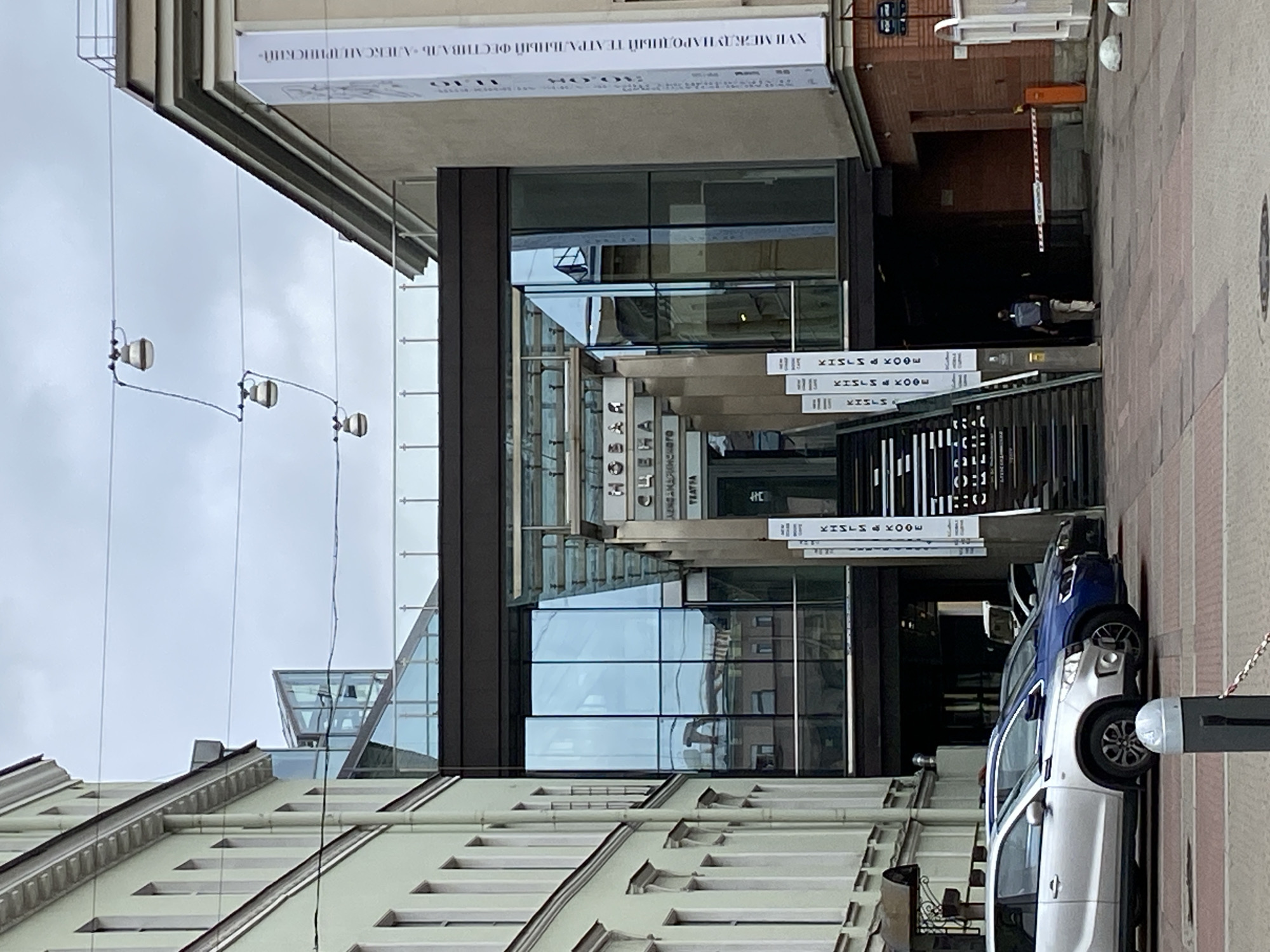 The image shows the exterior of a modern building complex with a glass facade and an elevated walkway connecting two structures. The building is located in Saint Petersburg, Russia. The architecture features a blend of classical and contemporary styles, with older buildings flanking a newer, sleek addition.
In the foreground, several cars are parked on a paved area. The building's entrance is visible, featuring a staircase leading up to the glass-enclosed section. Signs with Cyrillic text are displayed on white banners along the walkway and entrance. One prominent banner on the right side of the image appears to be advertising a cultural event.
Above the entrance, the word "НОВАЯ" (New) is prominently displayed in large letters, followed by "СЦЕНА" (Stage) in smaller lettering, and "АЛЕКСАНДРИНСКОГО ТЕАТРА" (Alexandrinsky Theatre). This indicates that the building is likely an extension or a separate venue associated with the Alexandrinsky Theatre.
The sky is overcast, suggesting a cloudy day. There are overhead wires with three decorative lamps suspended from them, adding to the urban aesthetic. A person wearing a backpack is walking away from the camera towards the entrance of the building. A red and white barrier gate is visible in the background.