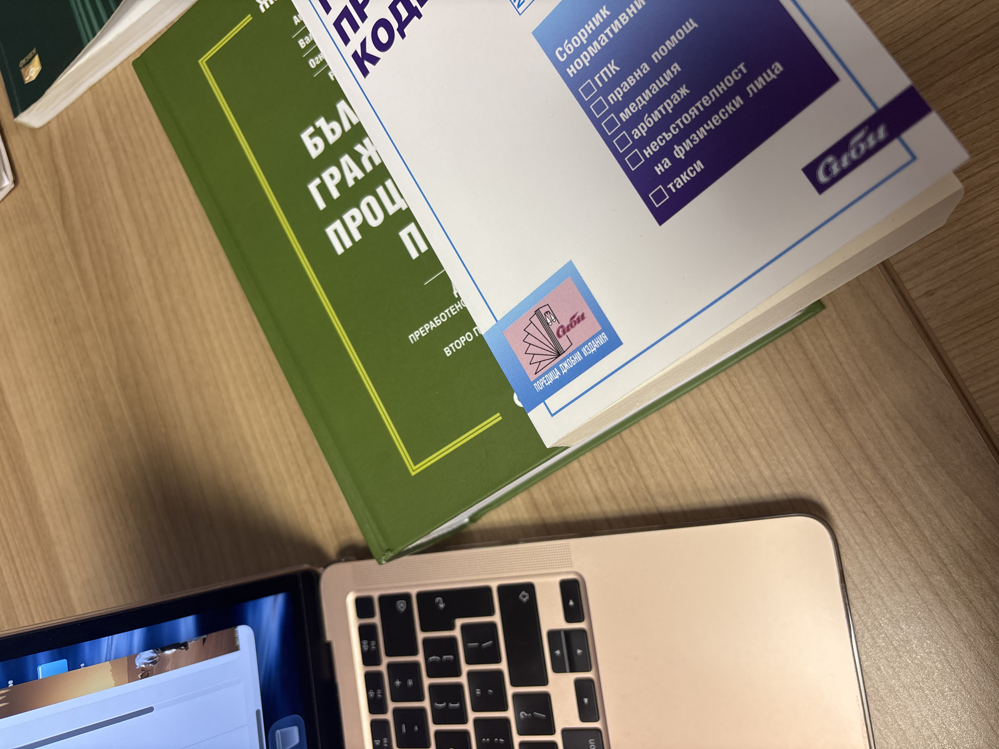 An overhead shot captures a study or work setup on a light-colored wooden desk. Two legal books and a partial view of a laptop are the main subjects.
A dark green hardbound book, likely a legal code, is positioned on the left, displaying its title "БЪЛ ГРАЖ ПРОЦ П" (likely Bulgarian Civil Procedure Code, as inferred from the OCR and common legal texts) and the text "ПРЕРАБОТЕНО ВТОРО П" (Revised Second Edition). To its right, a white paperback book titled "Сборник нормативни" (Collection of Normative Acts/Regulations) lists several legal topics: "ГПК" (Civil Procedure Code), "правна помощ" (legal aid), "медиация" (mediation), "арбитраж" (arbitration), "несъстоятелност на физически лица" (personal insolvency), and "такси" (fees). Both books feature the "Сиби" (Sibi) publisher logo, with the white book additionally stating "ПОРЕДИЦА ДЖОБНИ ИЗДАНИЯ" (Pocket Edition Series).
In the foreground, the lower right section of an open, gold-colored laptop (resembling a MacBook) is visible. Its keyboard, featuring a standard QWERTY layout with some Cyrillic characters, and a sliver of the display showing a graphical user interface, are discernible. The scene depicts an indoor setting, suggesting an environment for legal study or professional work. All visible text on the books is in Bulgarian.