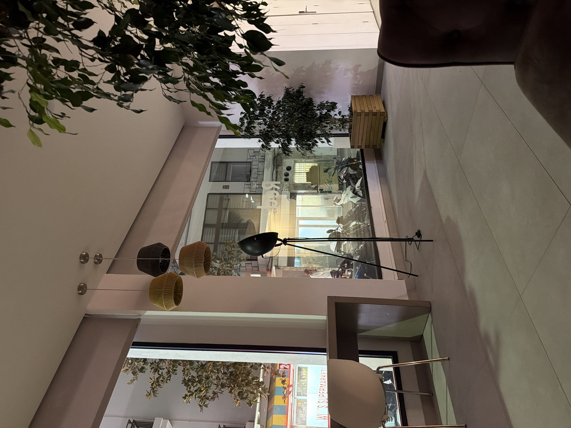 The image captures an indoor scene with a focus on decorative elements and architectural features. A large, leafy plant hangs from the ceiling on the left, its branches cascading downwards. Below this, a series of hanging light fixtures with woven, basket-like shades are visible. These fixtures are suspended by thin wires attached to the ceiling.
Further into the space, a floor lamp with a black shade is angled, casting a shadow on the wall. Behind this lamp, a large window or glass partition reveals the interior of another space, where motorcycles are parked. This area appears to be a commercial establishment, possibly a shop or showroom, given the presence of the vehicles. A wooden planter box with a plant sits on a ledge near the glass.
The flooring is tiled in a light, neutral color with subtle lines. In the lower part of the image, a glimpse of another area is visible through a doorway or opening. This area has a sign that reads "WI.S SUPERMARKET" in large letters. The overall impression is of a modern, perhaps retail or hospitality, interior with a mix of natural and artificial elements. The lighting suggests it is either daytime with artificial lights supplementing, or evening. The context of Sant Antoni De Portmany, Spain, places this scene within a tourist or urban environment.