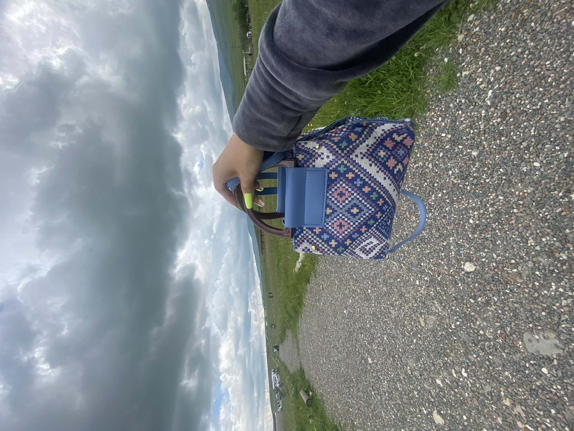 In Tsalkita, Russia, a person's hand, adorned with bright neon green fingernails, holds a small, intricately patterned handbag. The bag features a geometric design with blue, pink, and white diamond shapes, and has a solid blue flap closure. The person is wearing a dark gray velour jacket. They are standing on a gravel path that winds through a grassy field under a cloudy sky. In the background, a distant landscape features rolling green hills, some scattered buildings, and a few people walking in the distance. The overall scene suggests an outdoor setting in a rural or semi-rural area.