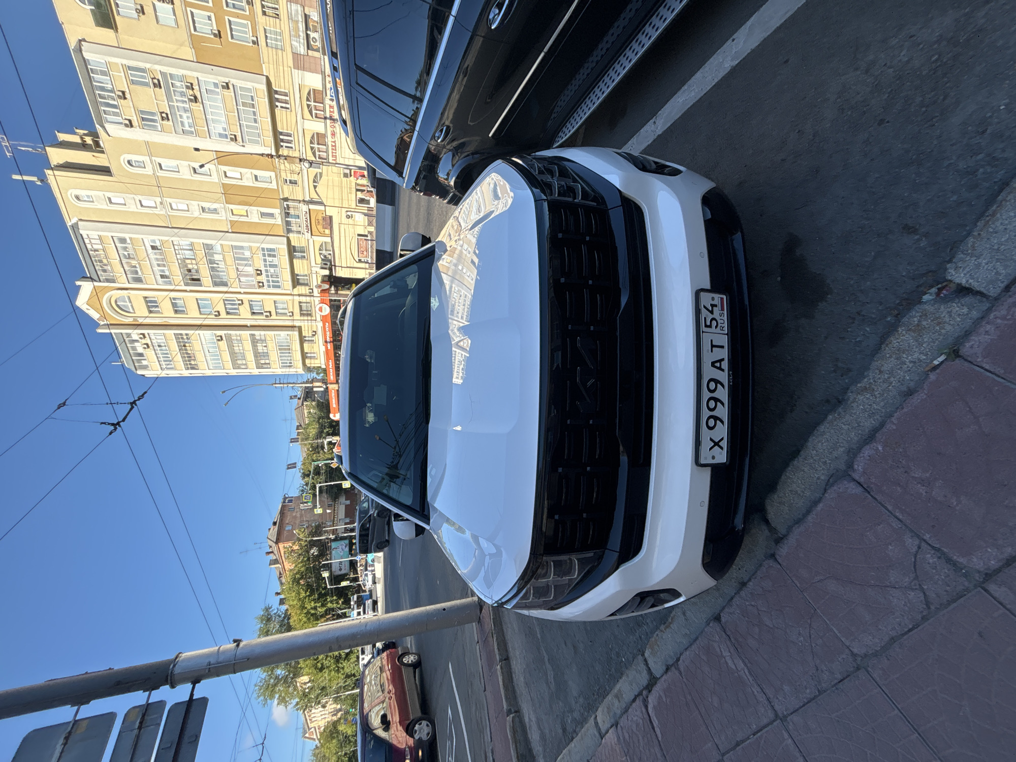 A white Kia Mohave SUV is parked on a city street in Novosibirsk, Russia. The car is facing the camera and is partially obscured by a dark SUV parked to its right. The license plate of the white Kia is visible and reads "X 999 AT 54 RUS". The street is paved with asphalt and has a sidewalk made of red tiles to the left of the white Kia. In the background, there are several buildings, including a tall, light-yellow apartment building with numerous windows and balconies, and other smaller buildings. The sky is clear and blue. Traffic lights and street signs are also visible, indicating an urban environment.