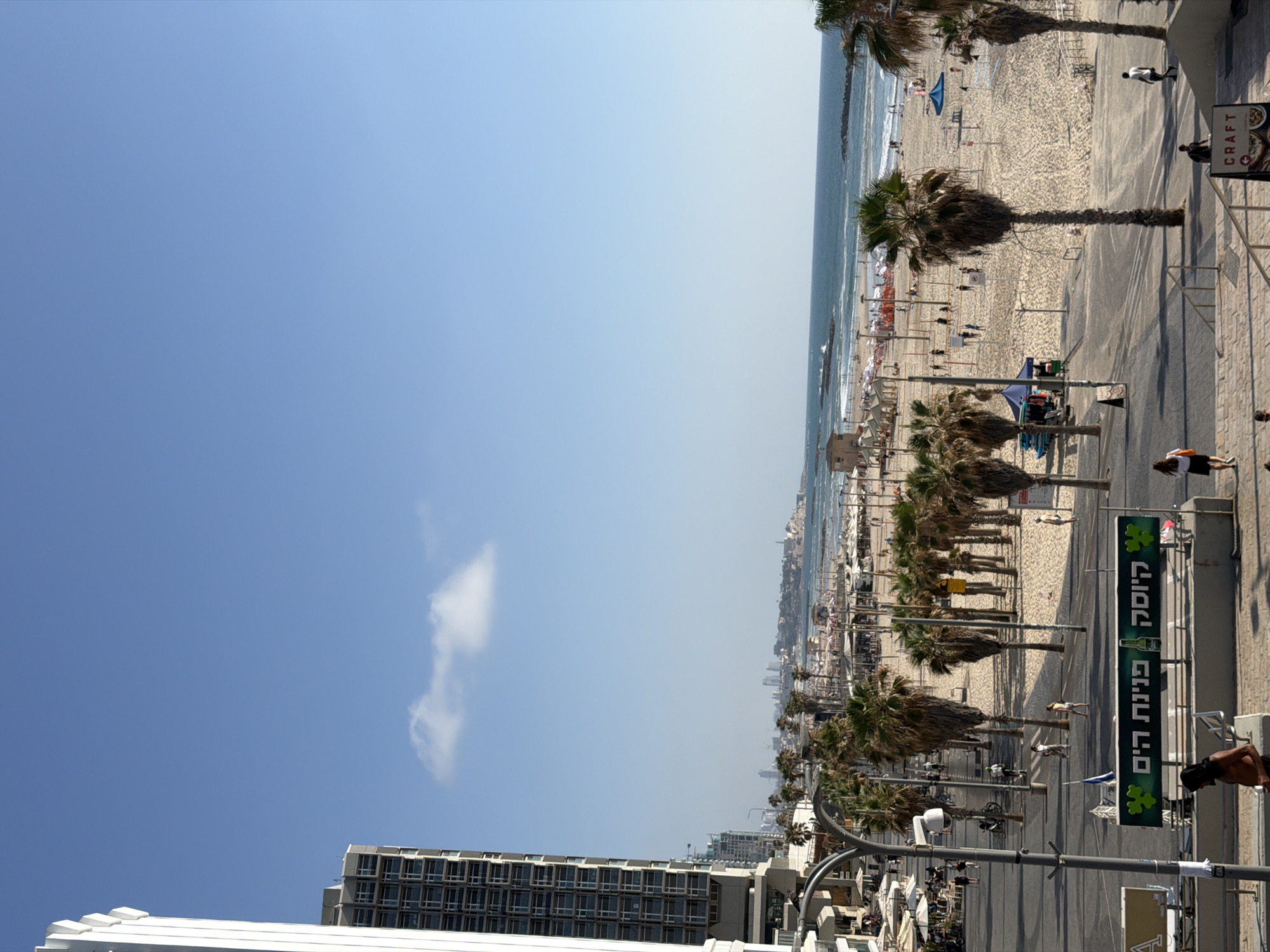 This is a daytime, eye-level, medium shot photograph taken from a slightly elevated vantage point overlooking a bustling beach and promenade in Tel Aviv, Israel. The scene captures a vibrant urban coastal environment.
In the foreground, a pedestrian walkway or stairs leads down towards a street. A large, green rectangular sign with Hebrew writing and clover leaf graphics is visible, with a bottle graphic next to the text "קיוסק". Several people are present on the walkway, including a bare-chested individual in the lower left. Further down the walkway, there are streetlights and parked bicycles.
Dominating the middle ground is a wide, sandy beach filled with people. Numerous beach umbrellas and lounge chairs are set up, suggesting a popular recreational area. Several palm trees are scattered across the beach and along the promenade. Beachgoers are seen walking, sitting, and playing beach sports like volleyball. A lifeguard tower is visible on the beach. The sea is calm with gentle waves, and a few scattered rocks are visible in the water.
In the background, the Tel Aviv skyline is visible. Tall, modern buildings, including a prominent white, multi-story structure on the left with many windows, are characteristic of the city's architecture. The sky is clear and blue with a few wispy clouds. The overall impression is one of a lively, sunny day at the beach in a major coastal city. The location appears to be near the Jaffa coastline, given the distant silhouette of historical buildings on the peninsula.