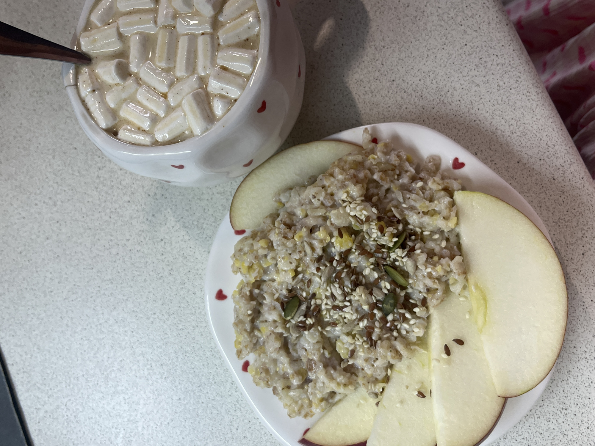 The scene, located in the city of Chaikovsky, Russia, presents a breakfast or snack arrangement on a light grey speckled countertop. A white ceramic plate with small red heart decals along its rim holds a serving of multi-grain porridge, possibly oatmeal, mixed with pale yellow kernels resembling corn. The porridge is topped with a sprinkle of brown flax seeds, white sesame seeds, and dark green pumpkin seeds. Alongside the porridge, several slices of a light-colored apple with reddish peel are arranged. To the right of the plate, a white ceramic mug, also adorned with small red heart decals, contains a creamy beverage topped with numerous miniature marshmallows and has a dark-colored spoon inserted. A dark object is partially visible in the upper left corner of the frame, and a pink and white patterned fabric item is partially visible in the bottom right corner.