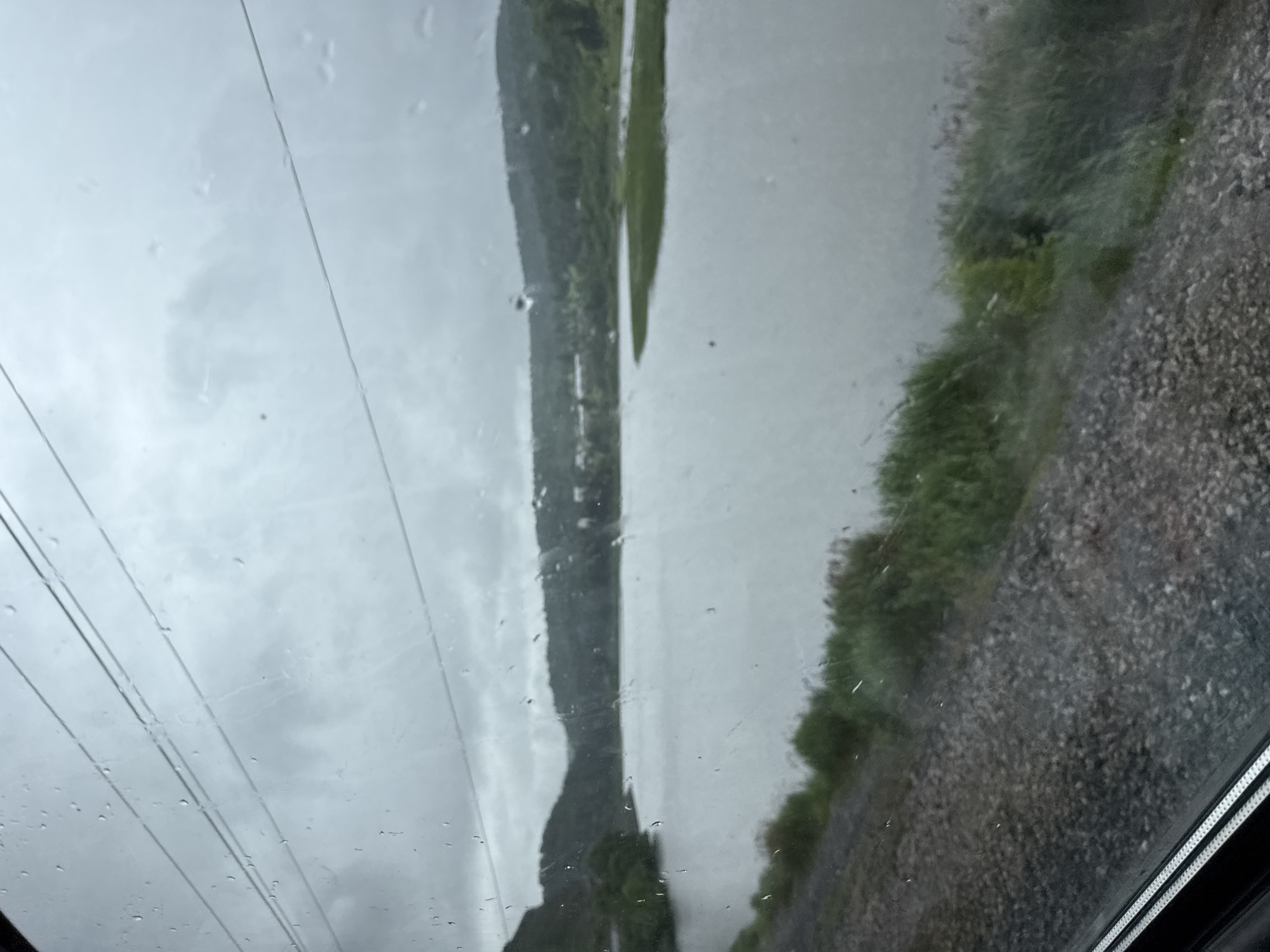 The image shows a body of water, likely a lake or river, viewed through a window with raindrops on it. The water is calm and reflects the overcast sky. Lush green vegetation lines the shore on the left side of the image, leading up to rolling hills covered in trees in the background. The sky is a uniform gray, indicating a cloudy or rainy day. In the foreground, a gravel embankment is visible, suggesting a railway track or road adjacent to the water. There are no people or specific identifiable landmarks visible in the image. The location is not specifically Minyar, Russia, as there are no visual cues to confirm this location.