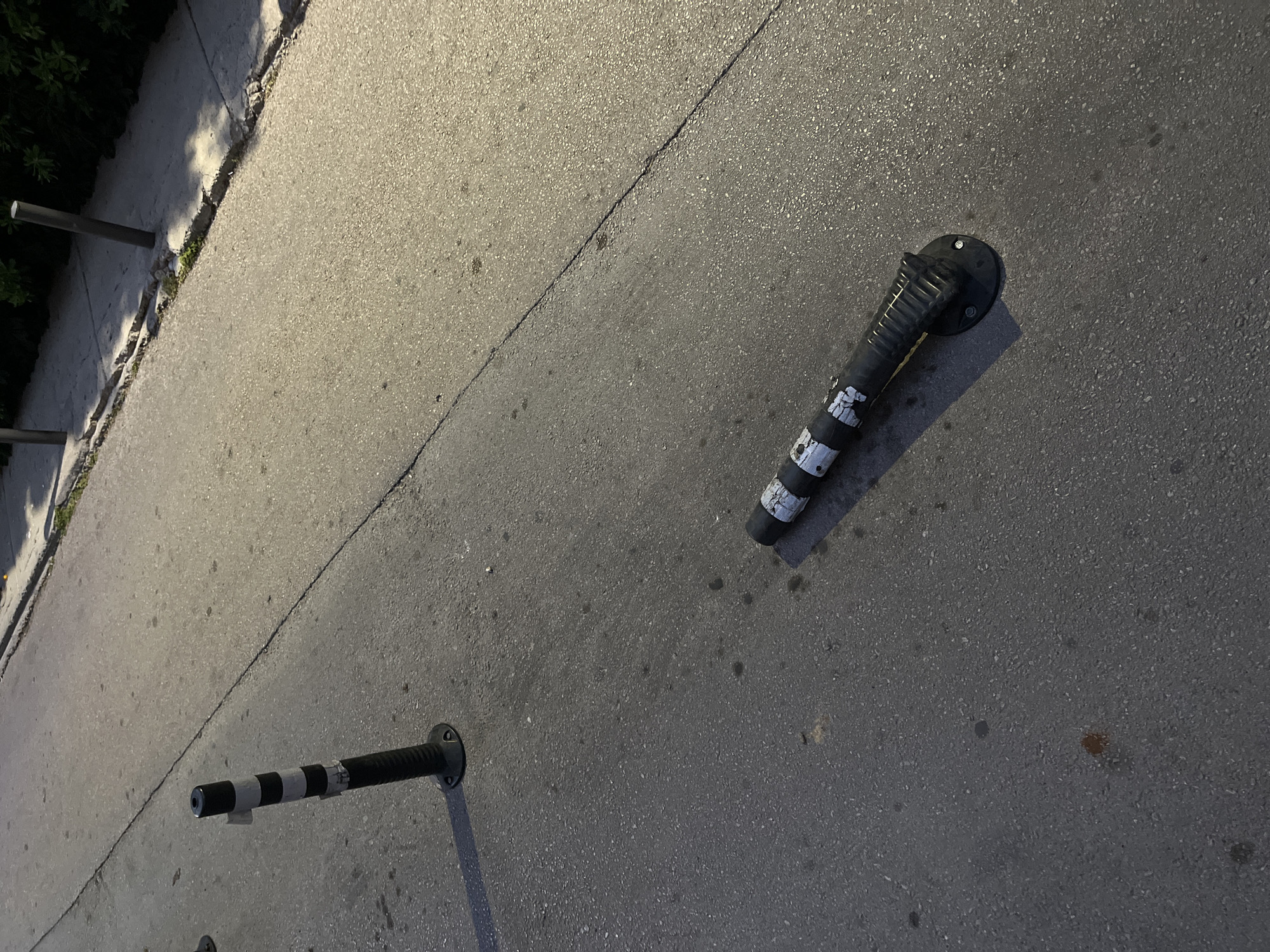 In Budva, Montenegro, two black and white striped traffic bollards are visible on an asphalt road. One bollard stands upright to the left of the frame, while the other lies horizontally on the ground to the right, its base plate exposed. The road surface shows some cracks and dark spots, possibly oil stains. The scene is illuminated by artificial light, casting shadows from the bollards. In the background, a concrete sidewalk with some vegetation and a few vertical poles is present. The overall impression is of a street environment, possibly near a parking area or roadway.