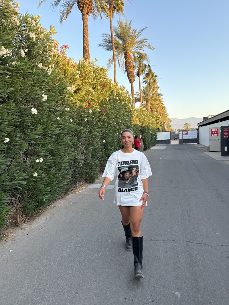A full shot captures a woman on a street in Indio, California. She is walking towards the camera, wearing a large white t-shirt with the words "TURBO BLANCO" and an image printed on it, along with black knee-high boots. A hedge, covered in white and red flowers, is to her left, and tall palm trees line the background. A white building with an emergency exit sign is visible behind her to the right. Other people are walking in the same direction, but further away, heading towards what appears to be an entrance area. The scene is bathed in bright sunlight, suggesting it is daytime.