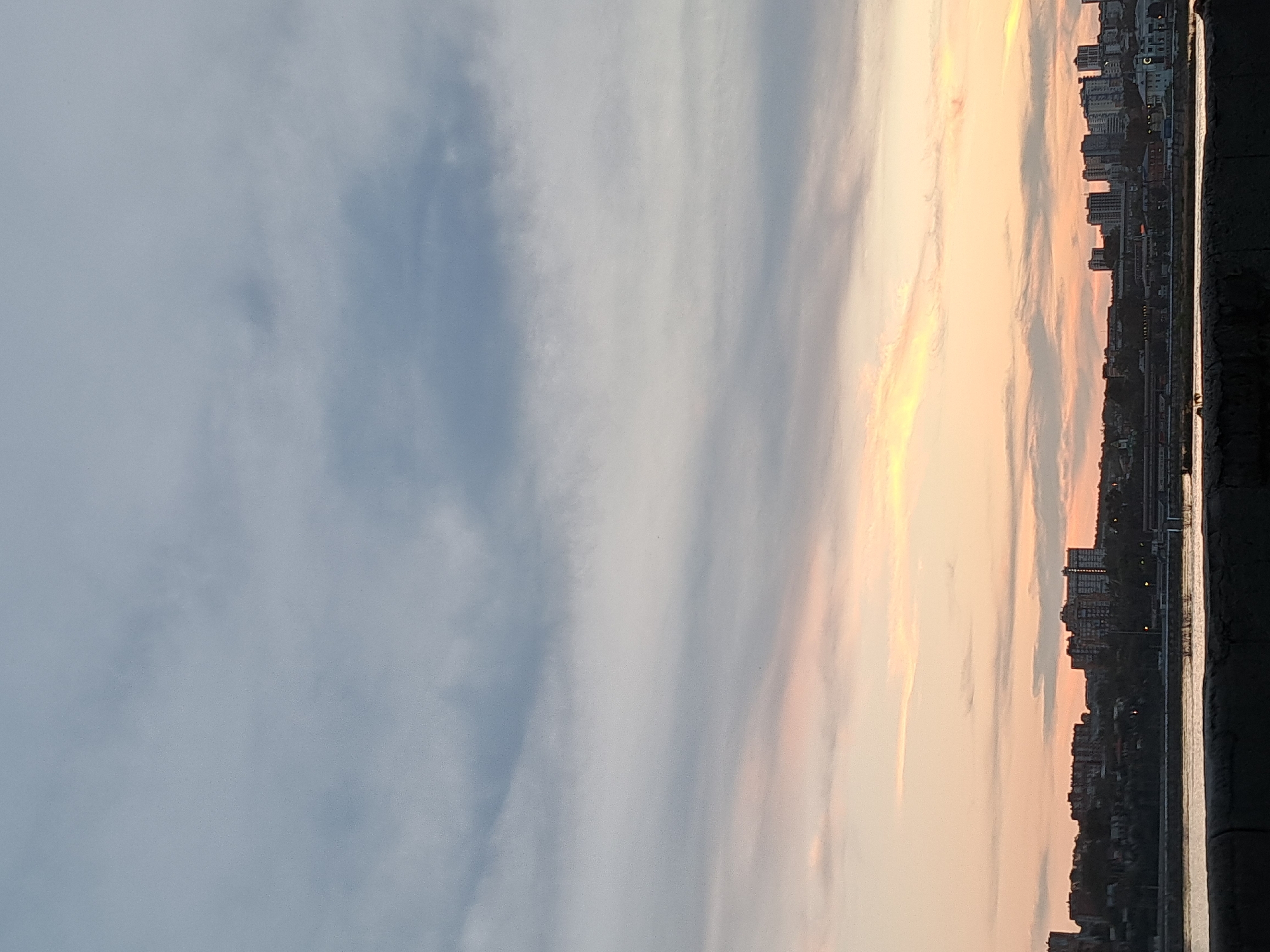 The image depicts a serene view of the Angara River in Irkutsk, Russia, during a twilight hour. The sky is a canvas of soft hues, transitioning from a pale grey at the top to delicate shades of pink and orange near the horizon. Wispy clouds stretch across the sky, catching the fading sunlight.
In the distance, the city skyline of Irkutsk is silhouetted against the colorful sky. Numerous buildings, ranging from apartment complexes to smaller structures, dot the landscape. The city appears to be situated on a gentle slope, with trees and vegetation interspersed among the buildings. A bridge is visible in the mid-ground, spanning the river and connecting different parts of the city.
The foreground of the image is dominated by the tranquil waters of the Angara River. The river's surface reflects the colors of the sky, creating a mirror-like effect. A dark, textured foreground element, possibly a retaining wall or embankment, runs along the bottom of the image, providing a sense of depth and perspective. The overall atmosphere is one of peace and quiet, characteristic of a city at dusk. There are no people or animals visible in the image.